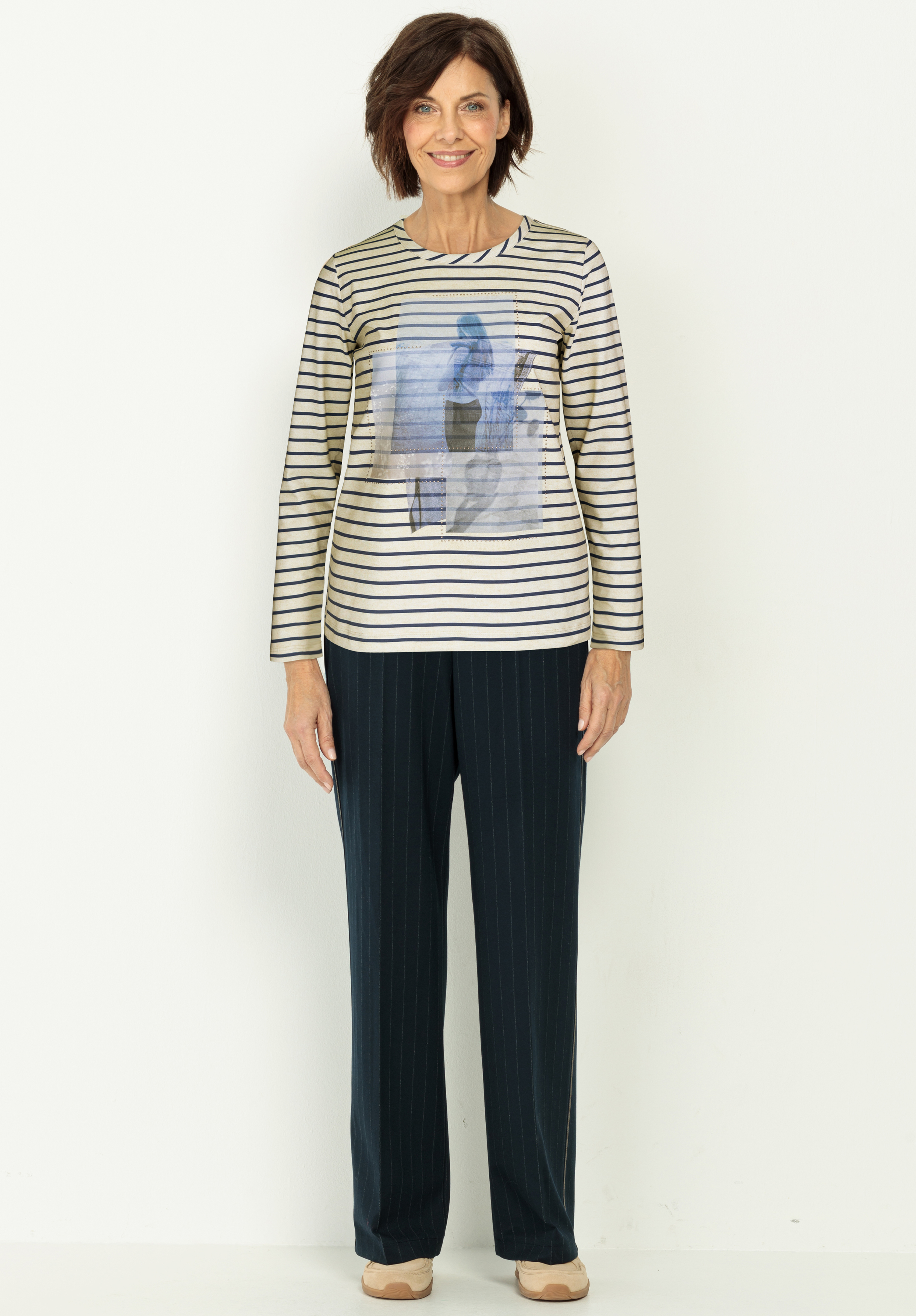 Eine Frau mit kurzen braunen Haaren lächelt, während sie vor einem weißen Hintergrund steht. Sie trägt ein gestreiftes langärmeliges Oberteil mit einem grafischen Muster und eine dunkle Nadelstreifenhose mit beigen Schuhen.