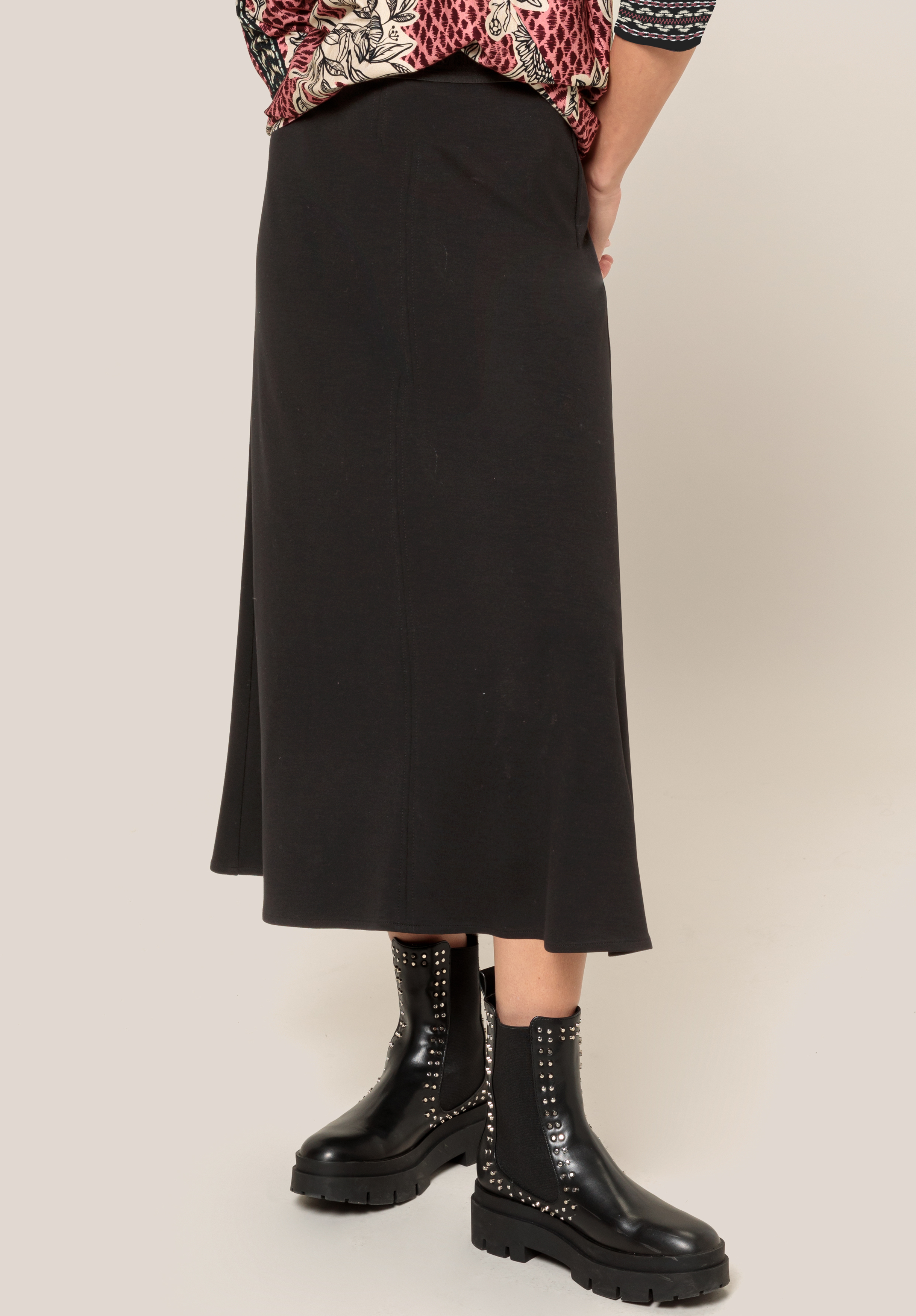 Eine Person in einem schwarzen Midirock, einem gemusterten Oberteil und schwarzen Stiefeletten mit Nieten steht vor einem schlichten Hintergrund. Das Foto ist von hinten aufgenommen und zeigt das Outfit von der Taille bis zu den Füßen.