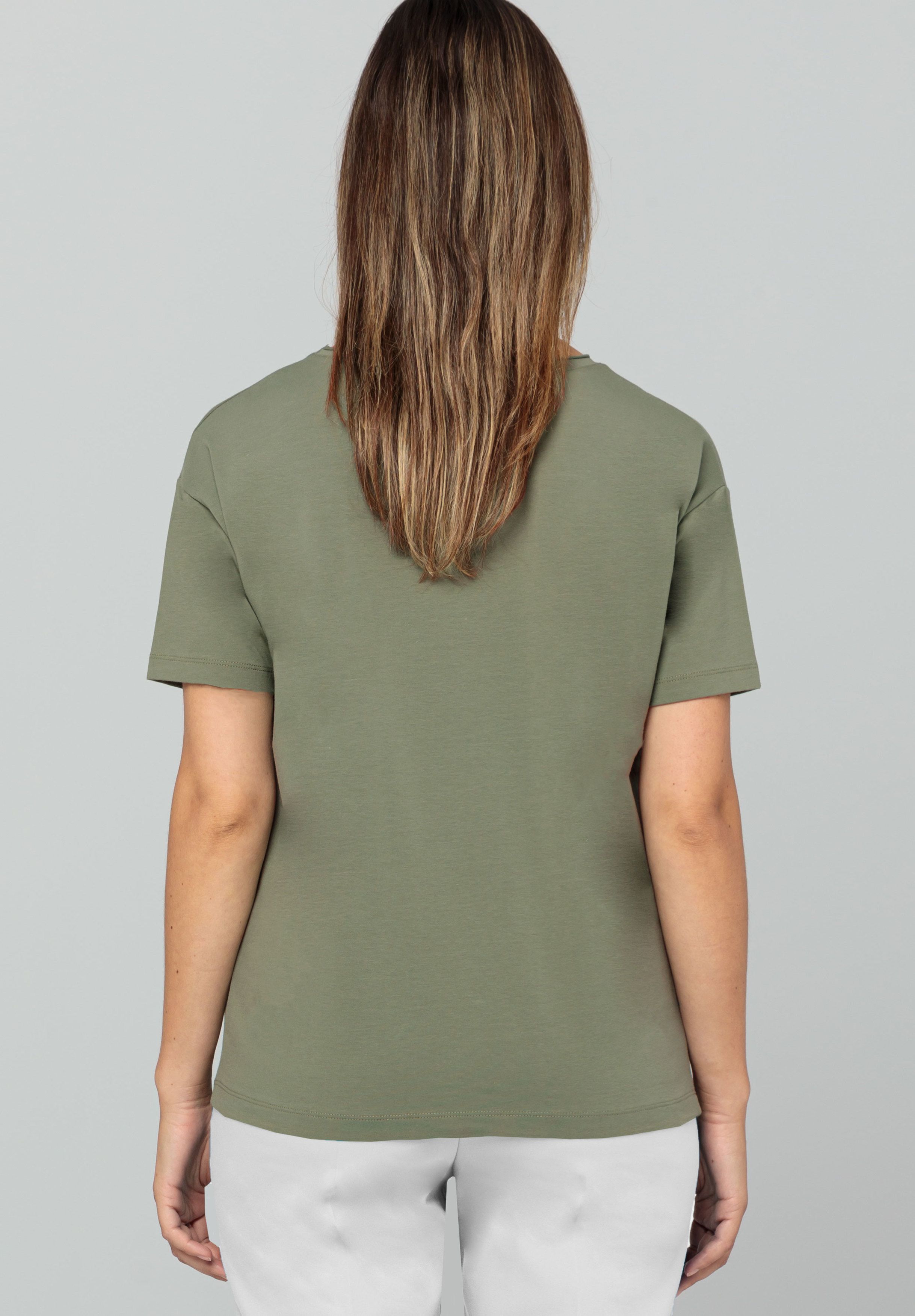 Eine Person mit mittellangem braunem Haar steht mit dem Rücken zur Kamera und trägt ein olivgrünes Kurzarmhemd und helle Hosen vor einem schlichten grauen Hintergrund.
