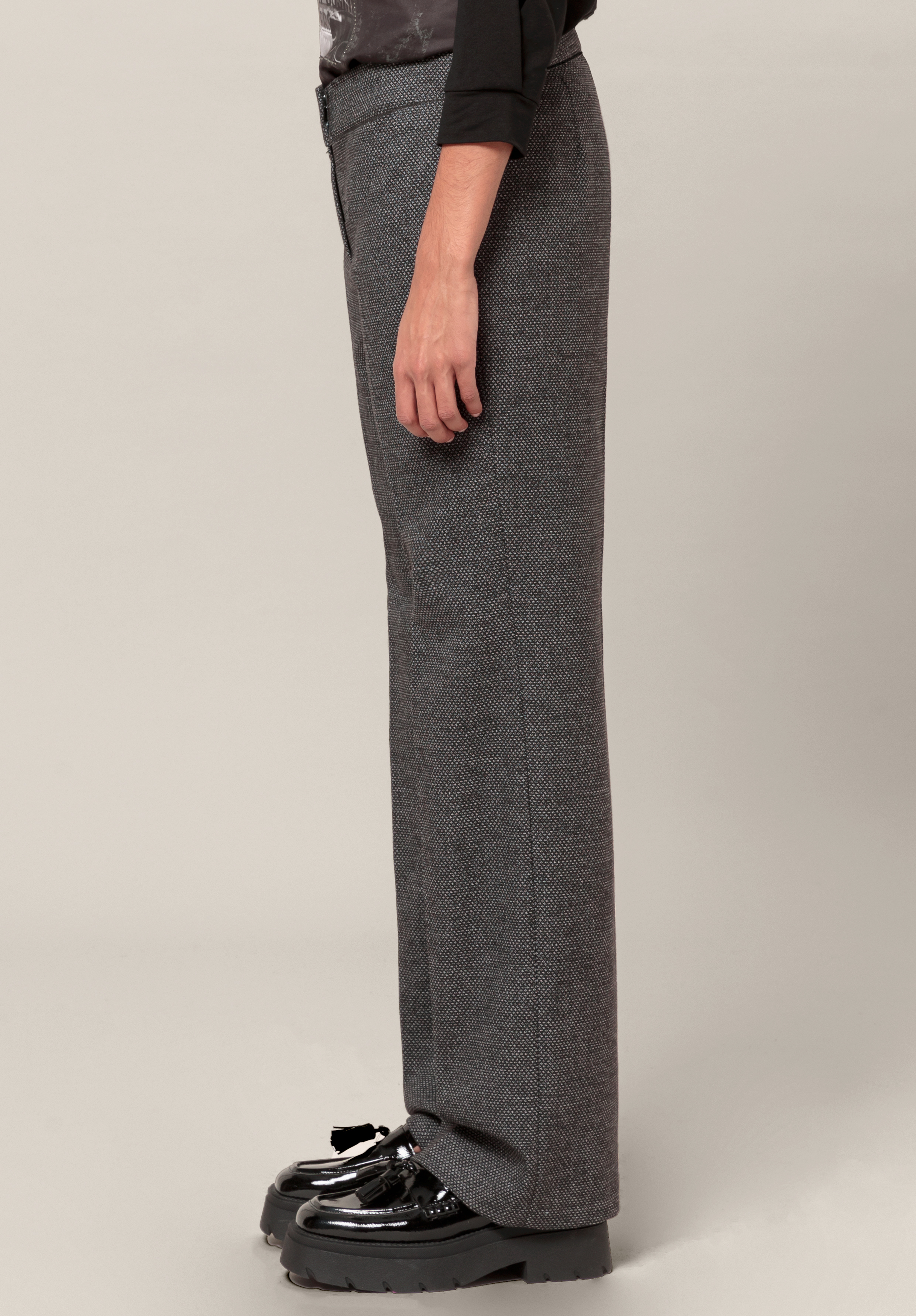 Eine Person steht seitlich und trägt eine graue Hose mit weitem Bein, ein schwarzes Langarmhemd und schwarze Plateauschuhe mit Schleifen vor einem schlichten hellen Hintergrund.
