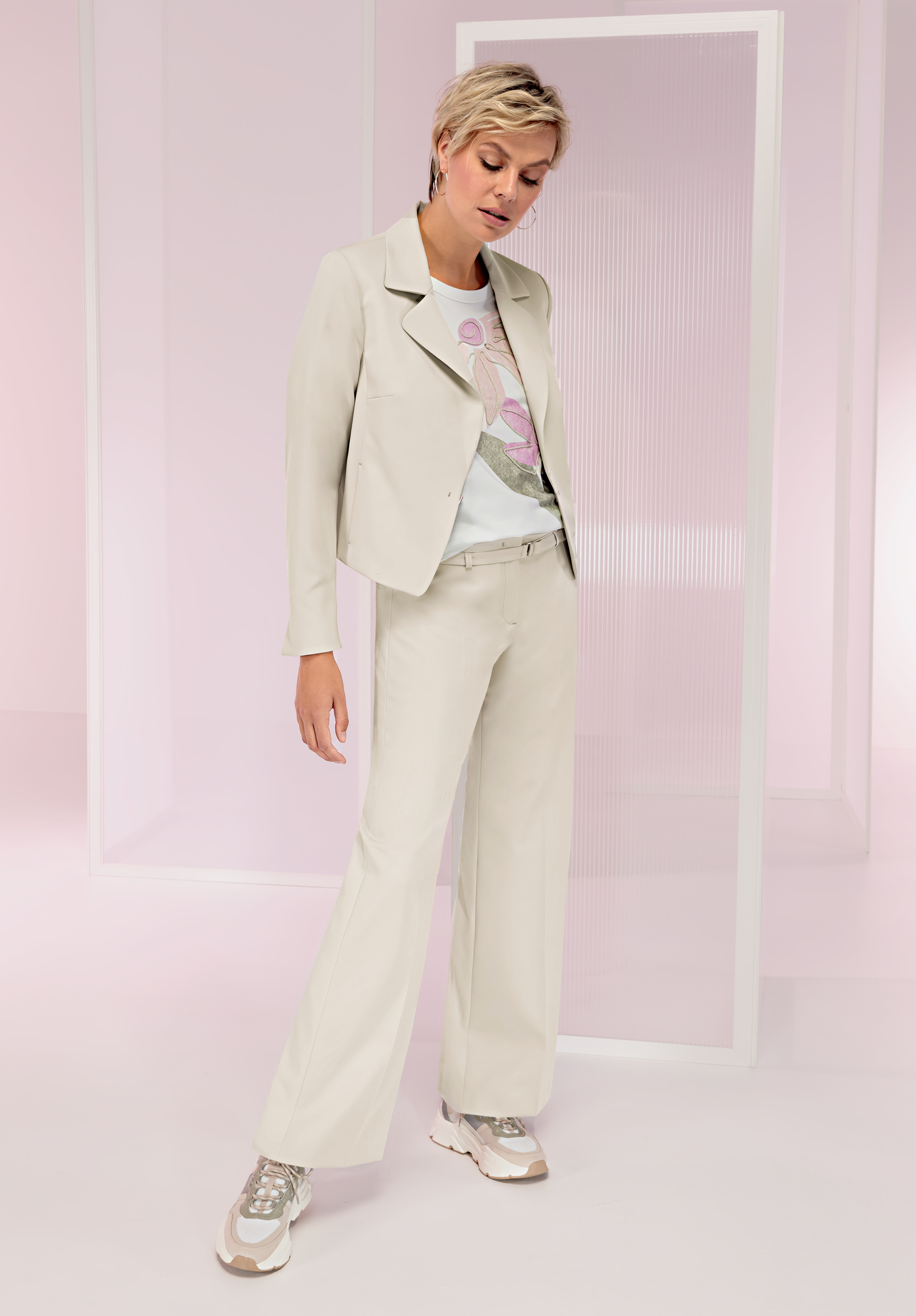 Eine Person mit kurzen blonden Haaren trägt einen hellen Anzug mit weiten Hosen, ein weißes T-Shirt mit Aufdruck und Turnschuhe und steht in einem modernen, sanft beleuchteten Raum mit durchscheinenden Trennwänden.