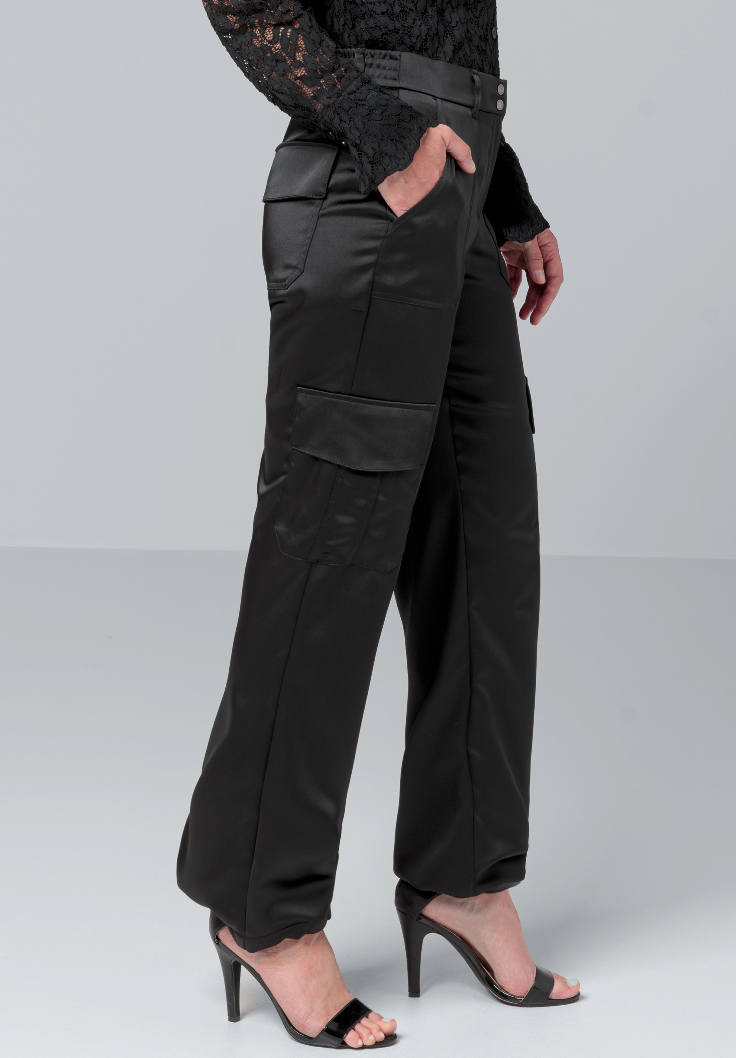 Eine Person trägt schwarze Cargohosen aus Satin mit Seitentaschen und ein langärmliges Oberteil aus schwarzer Spitze, dazu schwarze High Heels mit offenen Zehen, und steht mit einer Hand in der Tasche vor einem schlichten Hintergrund.