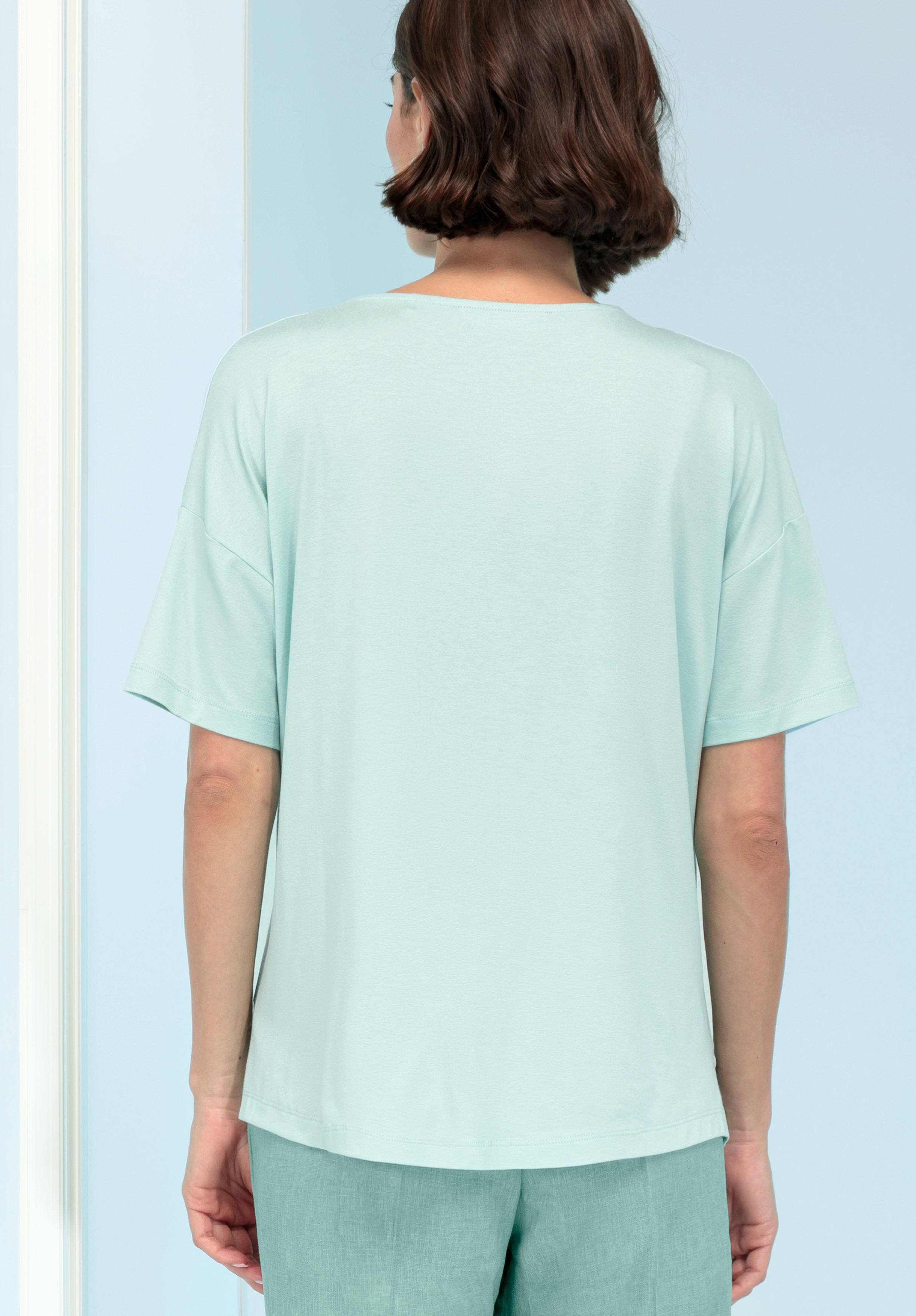 Von hinten ist eine Person mit kurzen braunen Haaren zu sehen, die ein locker sitzendes, hellmintgrünes Kurzarmoberteil und eine dazu passende Hose trägt und vor einem hellblauen Hintergrund steht.