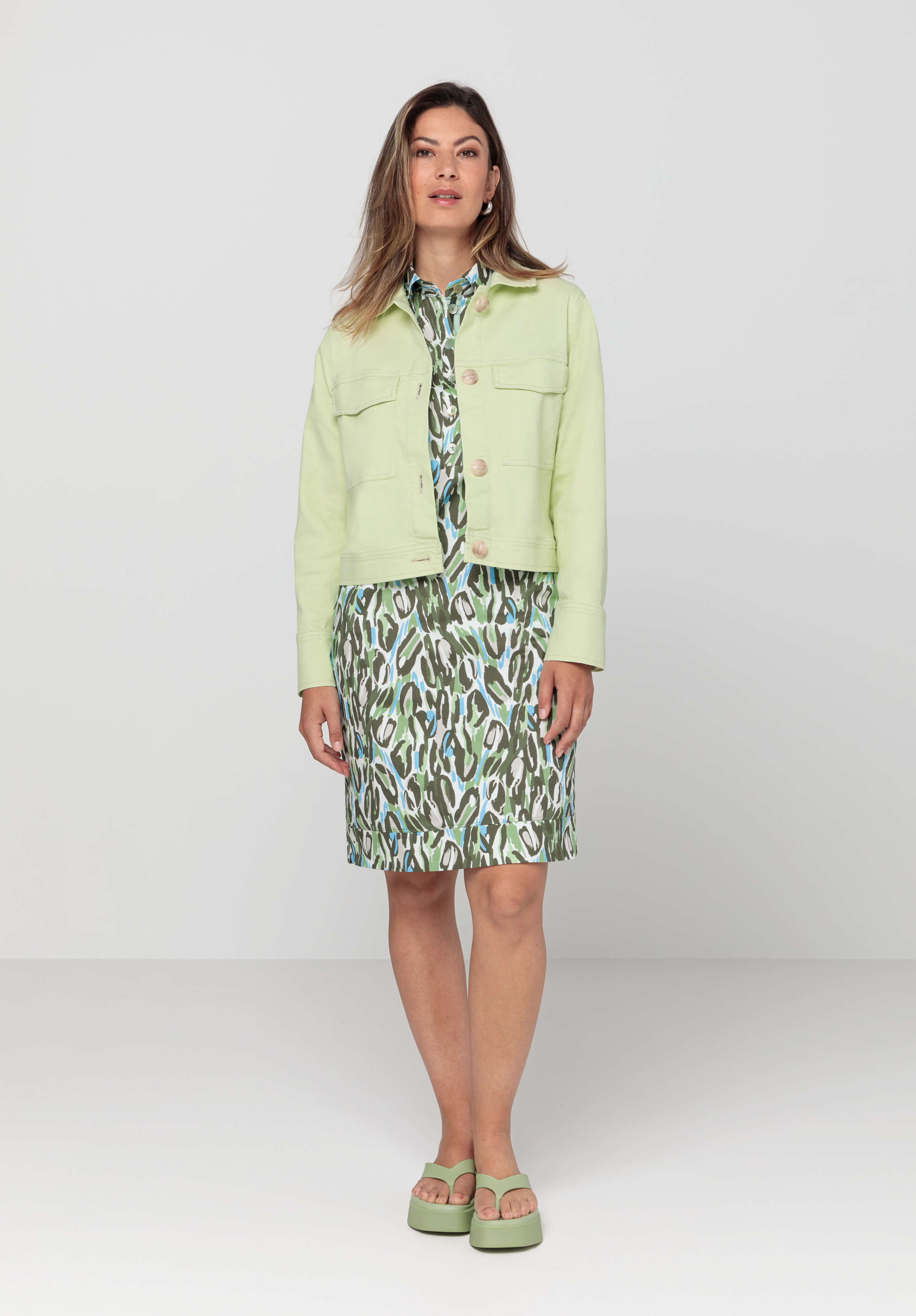 Eine Frau steht in einem Studio und trägt eine hellgrüne Kurzjacke über einem grün-weiß gemusterten Kleid mit passenden Sandalen. Ihr Blick ist nach vorne gerichtet, ihr Gesichtsausdruck neutral, vor einem schlichten weißen Hintergrund.
