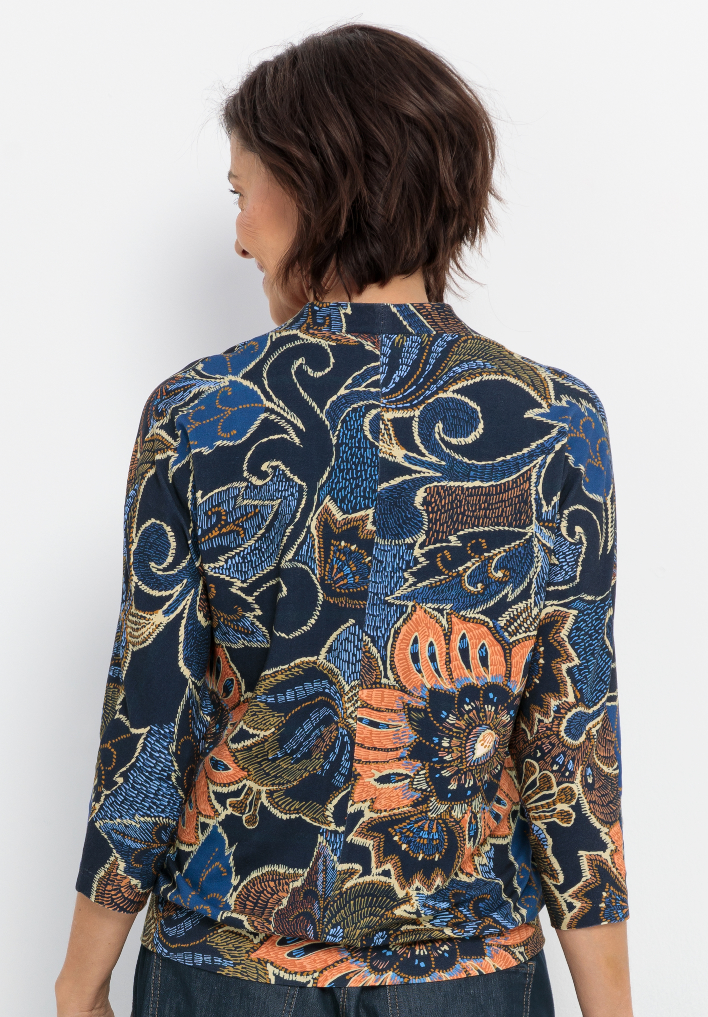 Eine Frau mit kurzen braunen Haaren, von hinten gesehen, trägt ein marineblaues Oberteil mit einem auffälligen, mehrfarbigen Blumen- und Paisleymuster in Orange, Gold und Blau. Das Oberteil hat dreiviertellange Ärmel.