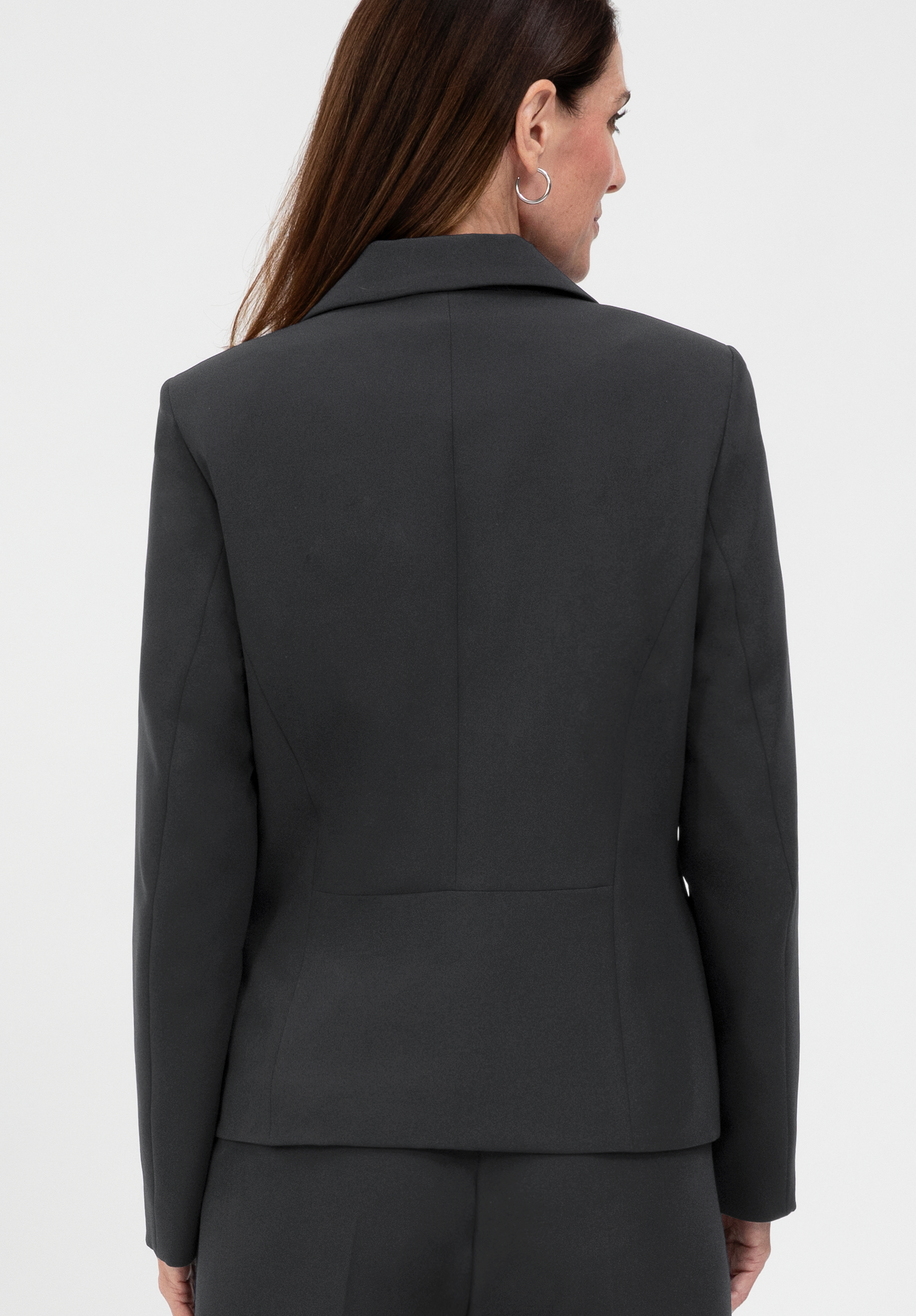 Vor einem schlichten weißen Hintergrund ist eine Frau mit langen braunen Haaren von hinten zu sehen, die einen dunkelgrauen, taillierten Blazer und eine dazu passende Hose trägt.