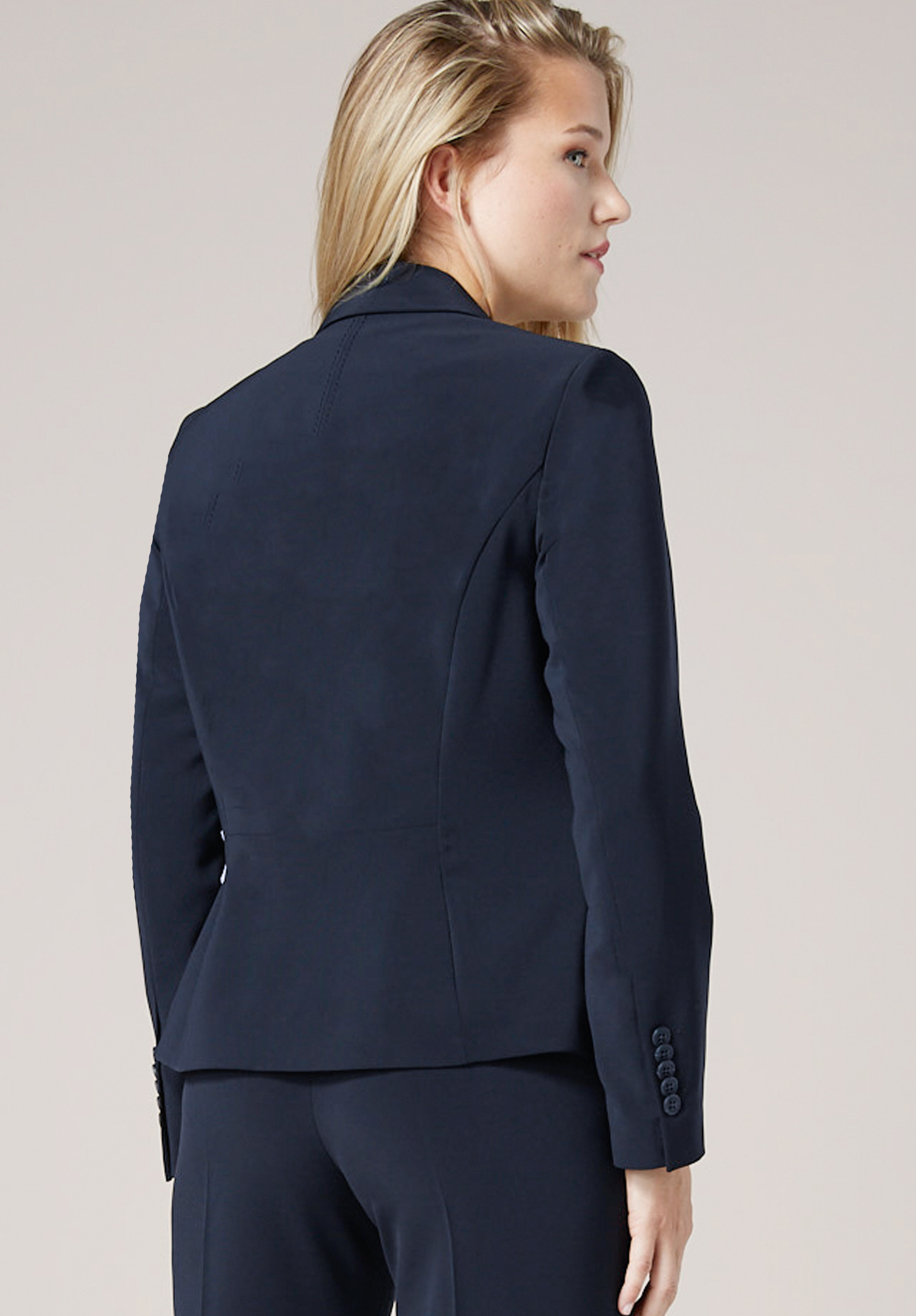 Eine Frau mit langen blonden Haaren ist von hinten zu sehen. Sie trägt einen dunklen marineblauen Blazer und eine passende Hose und steht vor einem schlichten hellen Hintergrund.