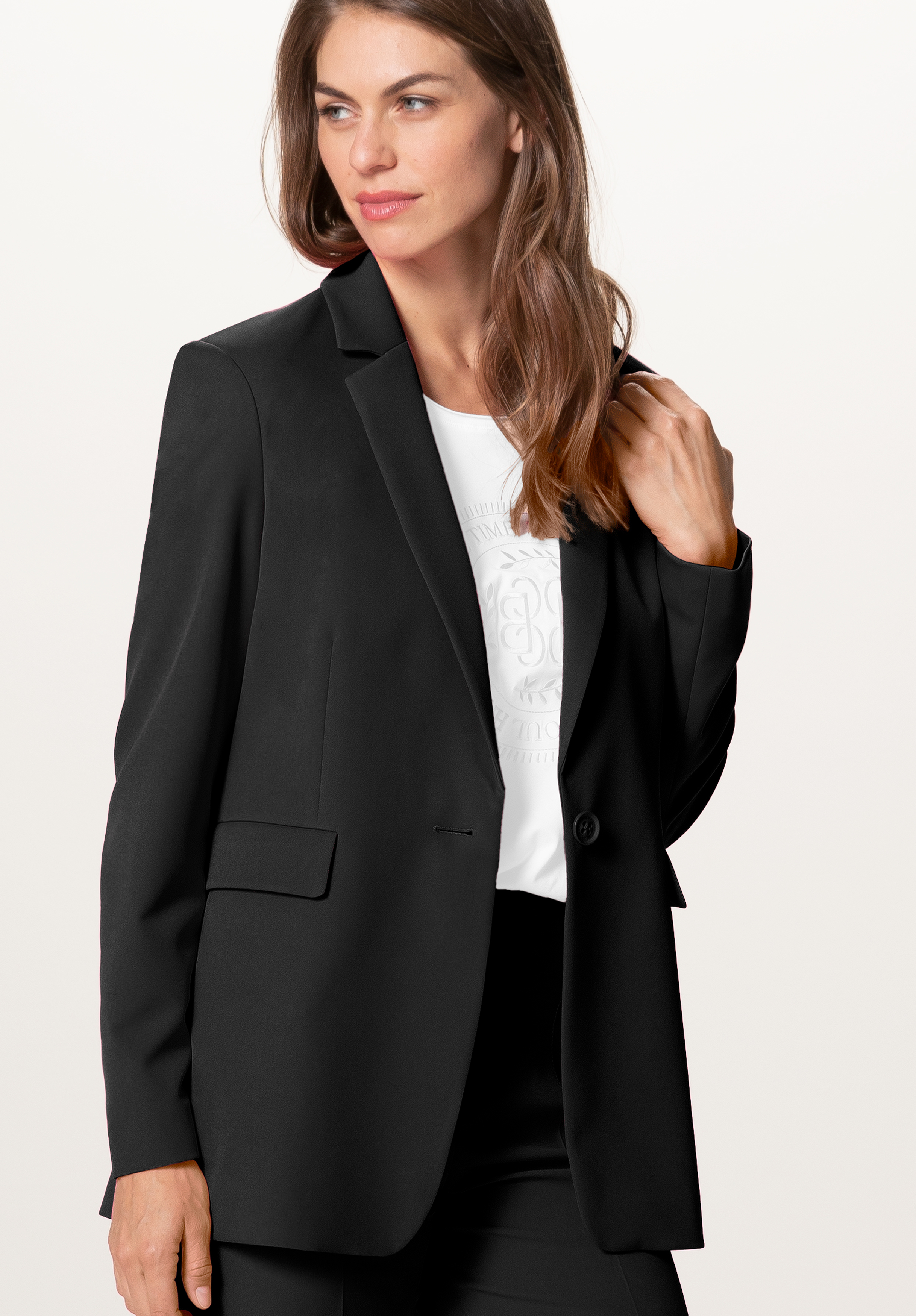 Eine Frau mit langen braunen Haaren trägt einen schwarzen Blazer über einer weißen Bluse mit dezentem Muster; sie steht vor einem schlichten hellen Hintergrund und blickt mit einem leichten Lächeln zur Seite.