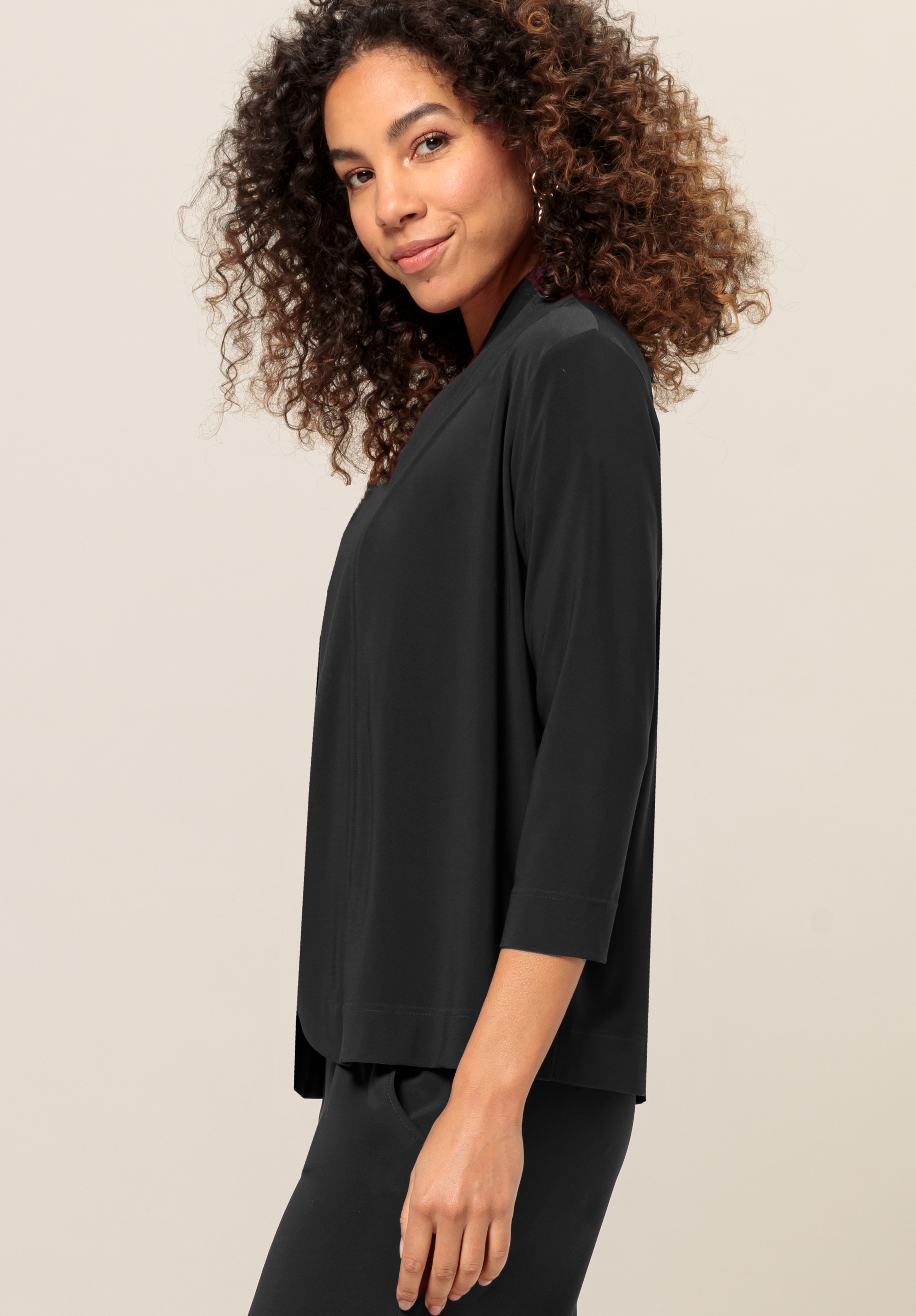 Eine Frau mit lockigem Haar, die eine schwarze Bluse mit Dreiviertelärmeln und eine dazu passende schwarze Hose trägt, steht seitlich und lächelt leicht vor einem neutralen Hintergrund.