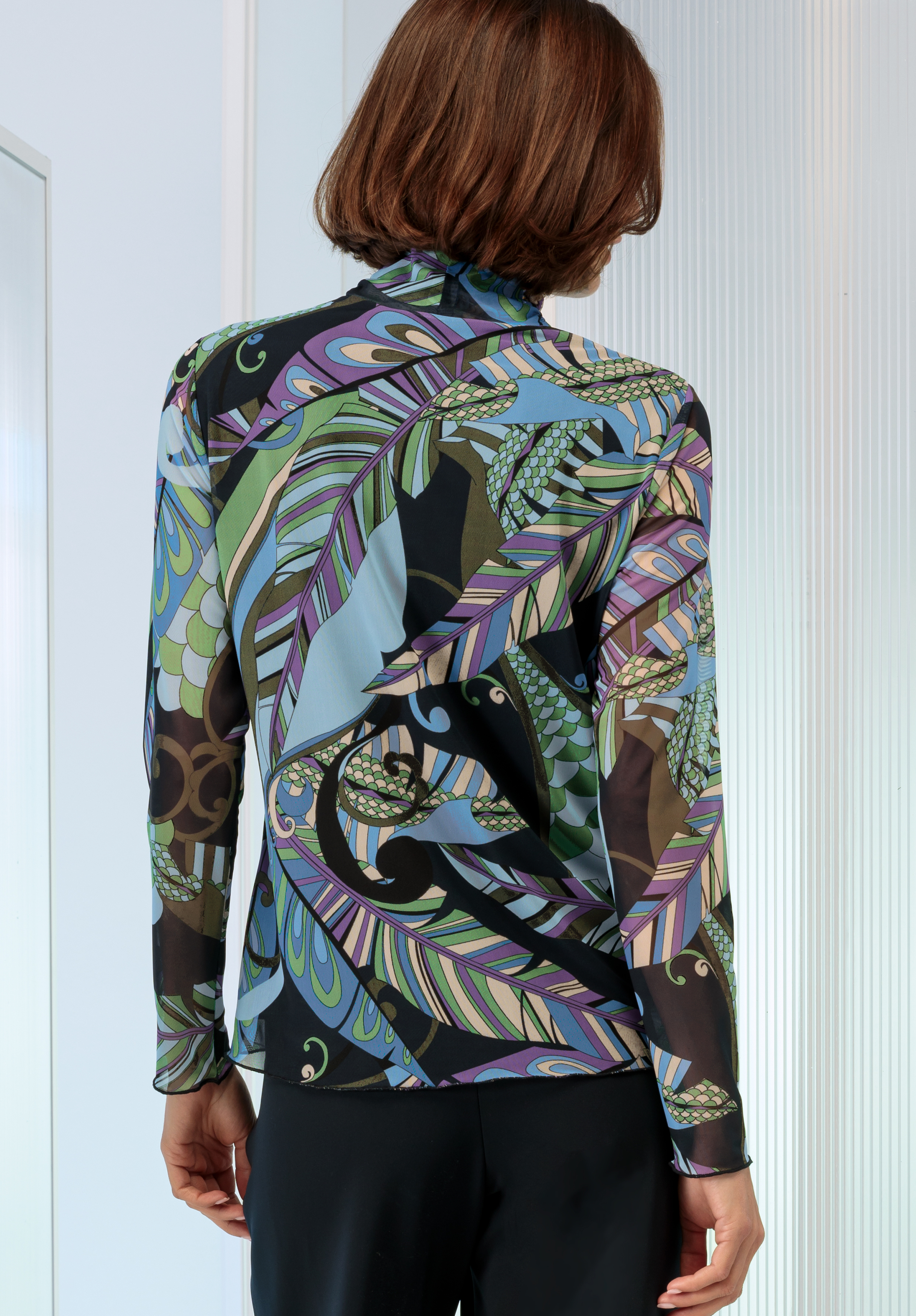 Eine Person mit kurzen braunen Haaren trägt von hinten gesehen ein langärmliges, hochgeschlossenes Oberteil mit einem farbenfrohen, abstrakten geometrischen Blattmuster in den Farben Lila, Blau, Grün und Schwarz.