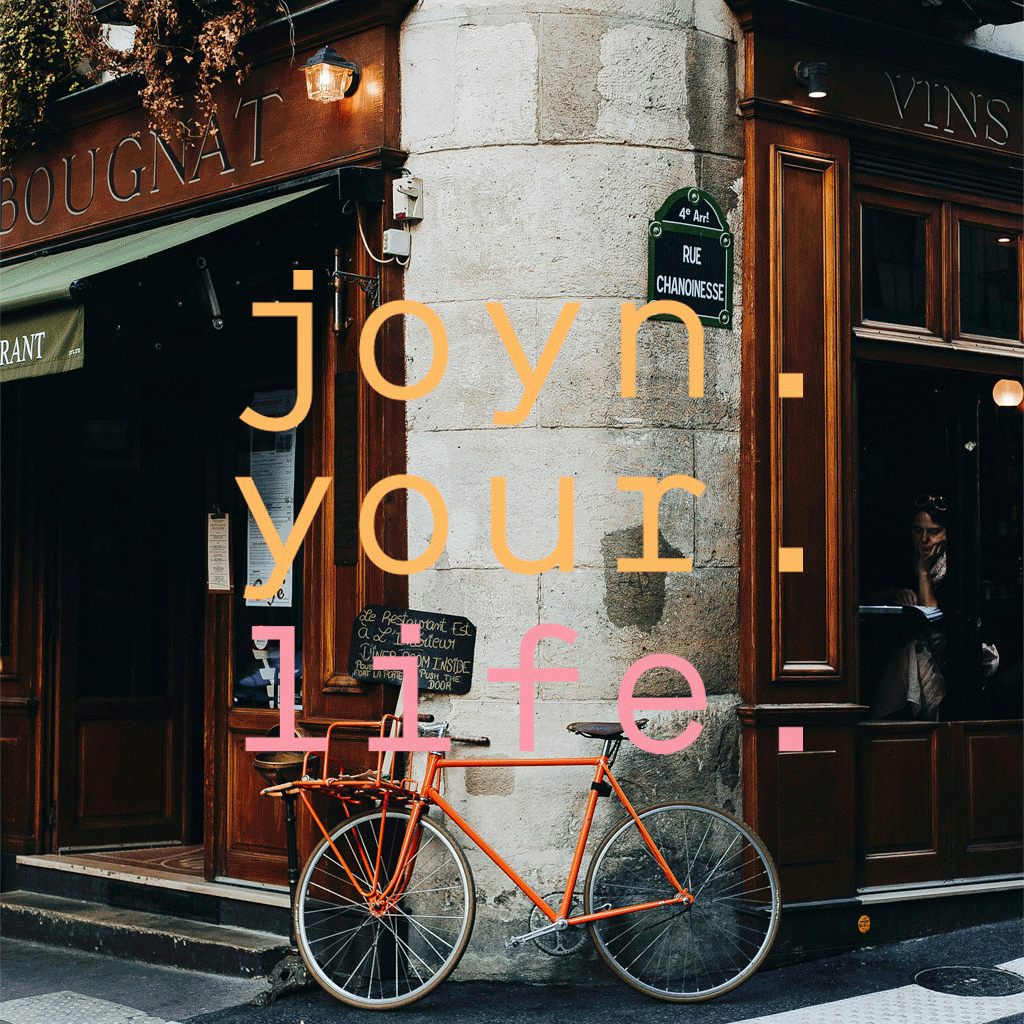Ein orangefarbenes Fahrrad ist neben einem Eckcafé mit hölzernem Äußeren und grünen Schildern geparkt. Der überlagerte bunte Text lautet: "joyn: your l1fe.".