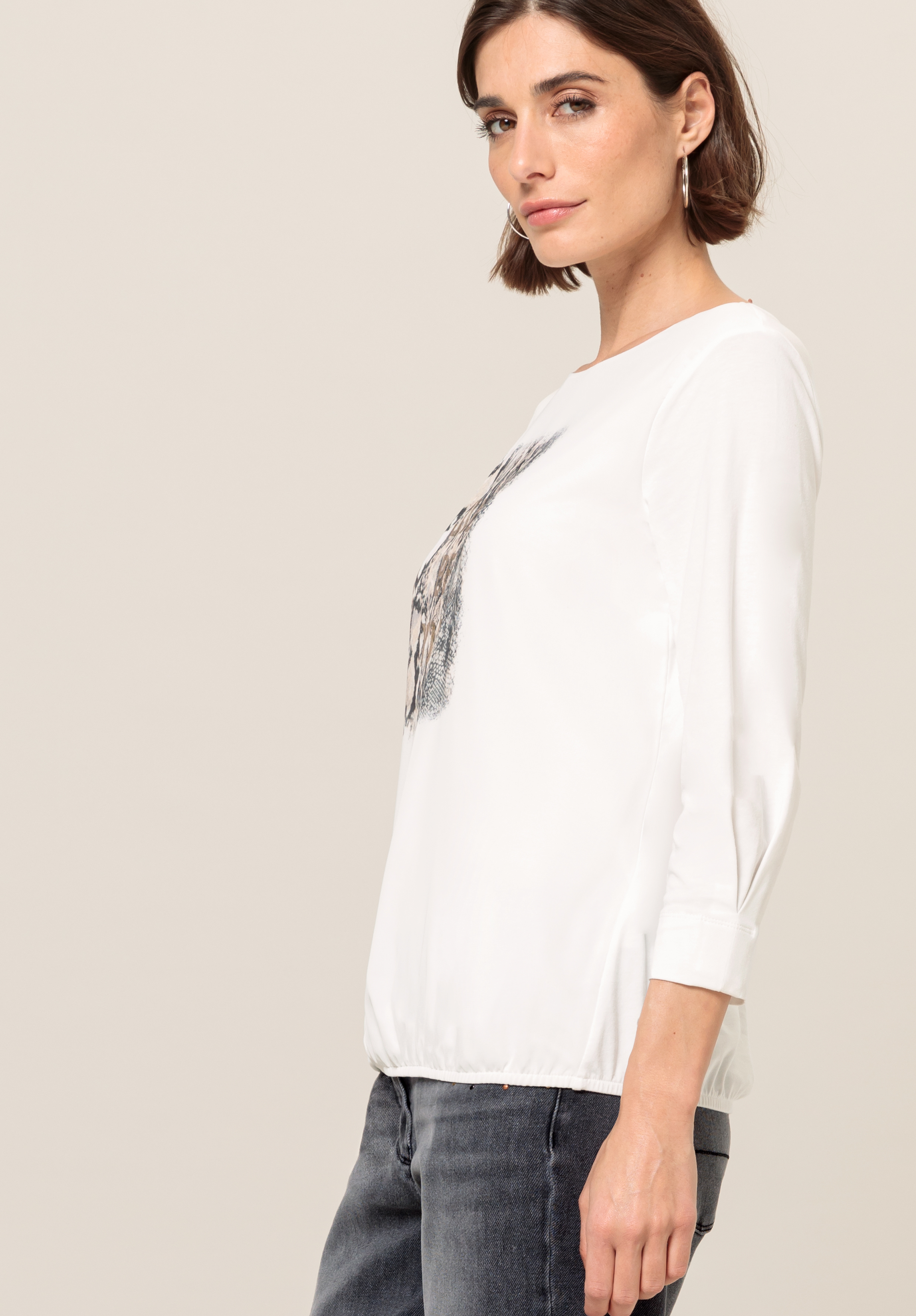 Frau mit kurzen braunen Haaren, die ein weißes Langarmshirt mit dezenter Grafik und graue Jeans trägt und im Profil vor einem schlichten hellen Hintergrund steht.