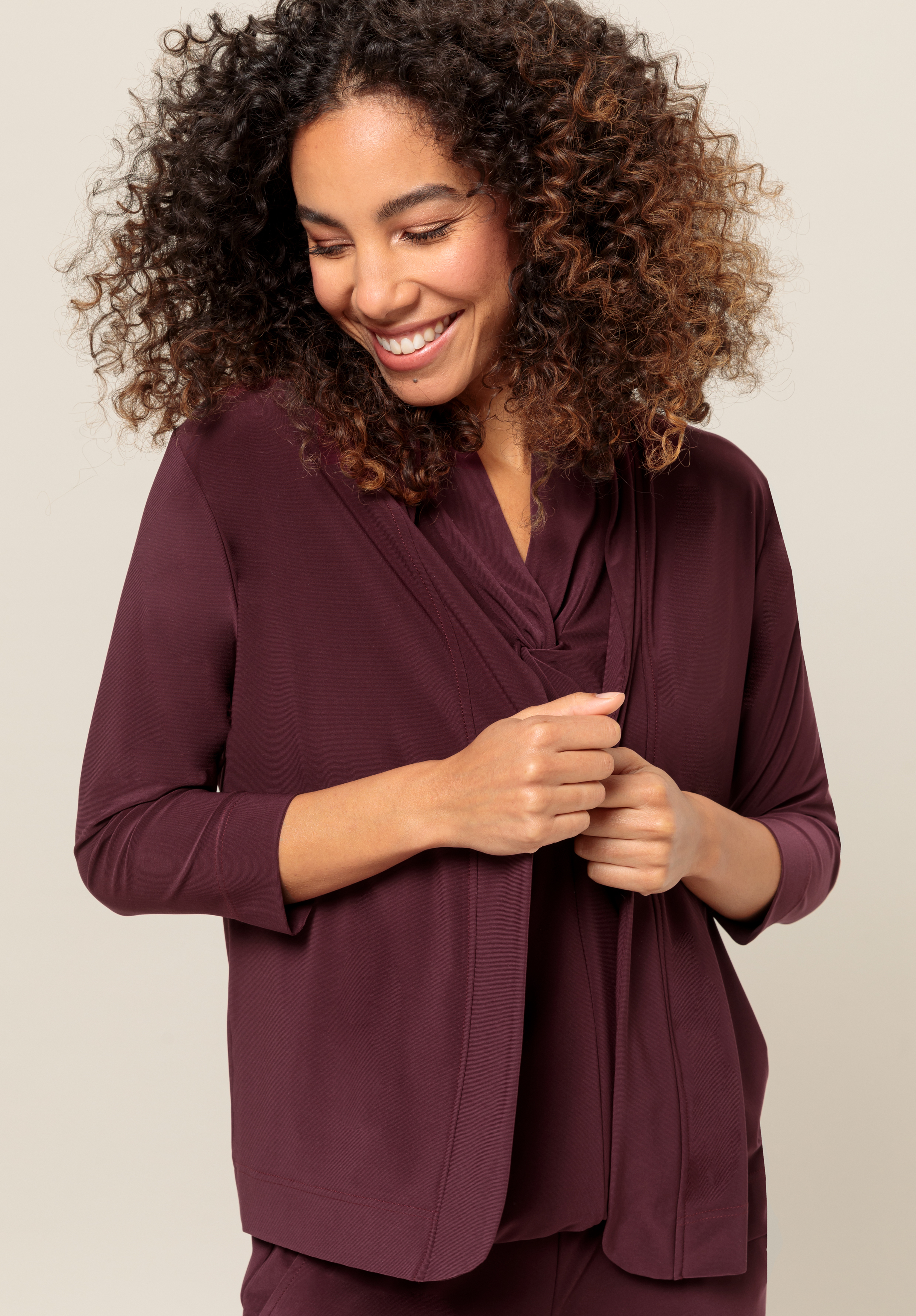 Eine Frau mit lockigem Haar lächelt und schaut nach unten, während sie einen burgunderfarbenen Blazer über einem passenden Oberteil trägt und vor einem neutralen Hintergrund steht.