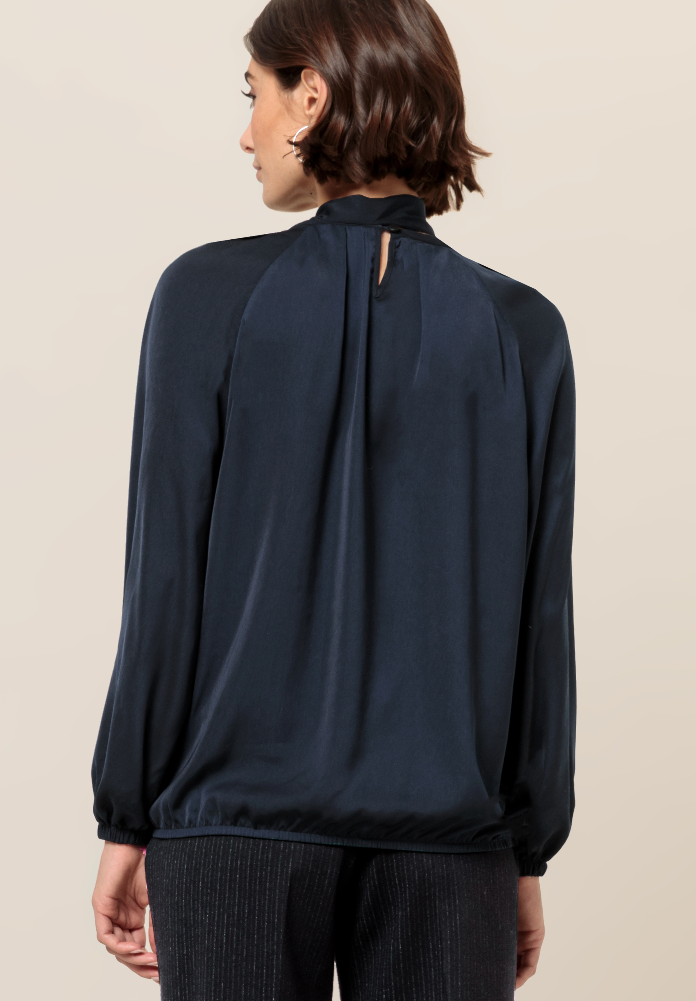 Eine Frau mit kurzen braunen Haaren ist von hinten zu sehen. Sie trägt eine langärmelige, dunkelblaue Bluse mit hohem Kragen und geknöpftem Schlüssellochdetail. Dazu trägt sie eine dunkle, strukturierte Hose.