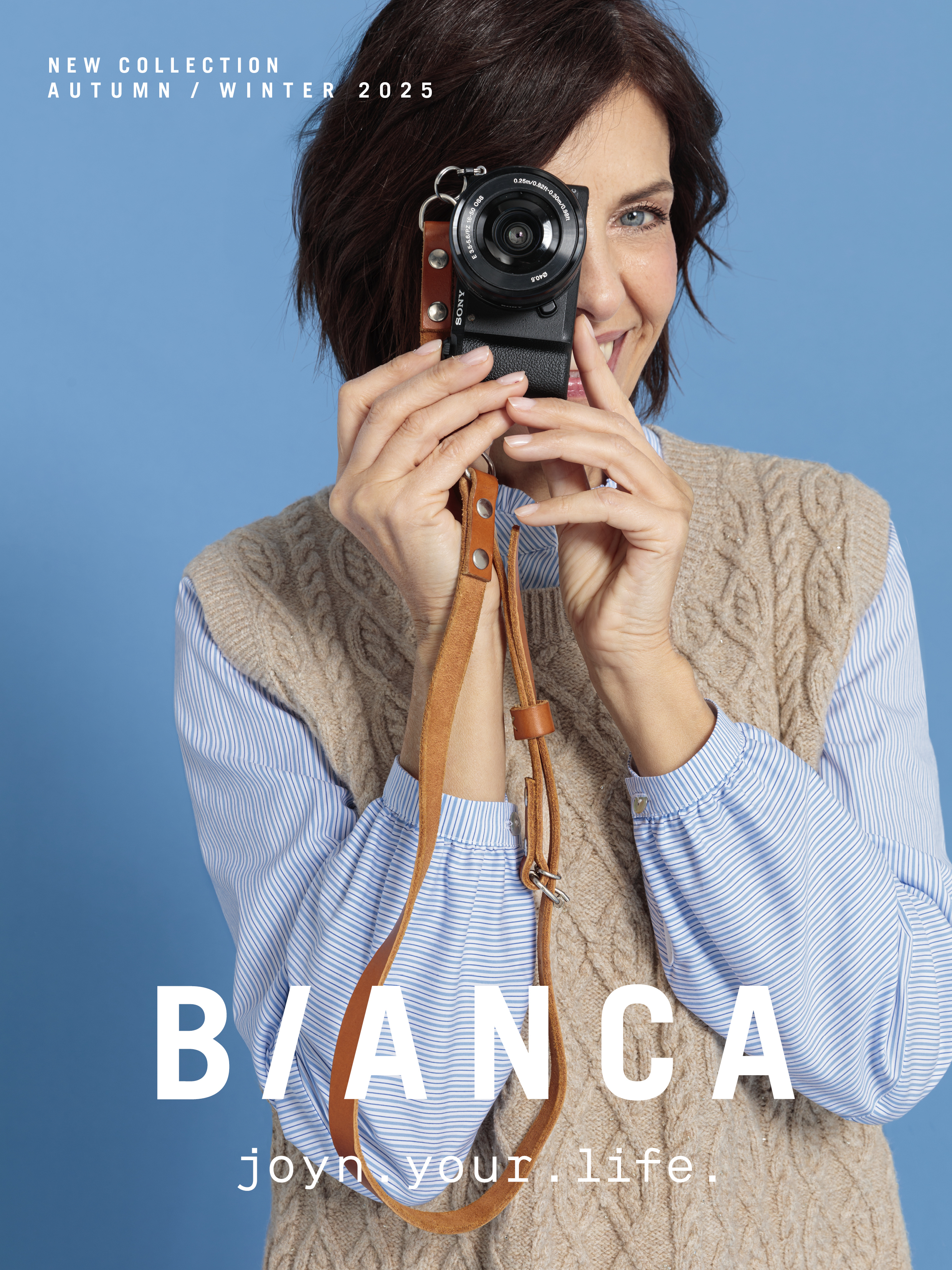 Eine lächelnde Frau in Zopfstrickweste und gestreiftem Hemd hält vor einem blauen Hintergrund eine Kamera an ihr Gesicht. Der Text lautet: Neue Kollektion Herbst/Winter 2025. BIANCA. joyn your life.