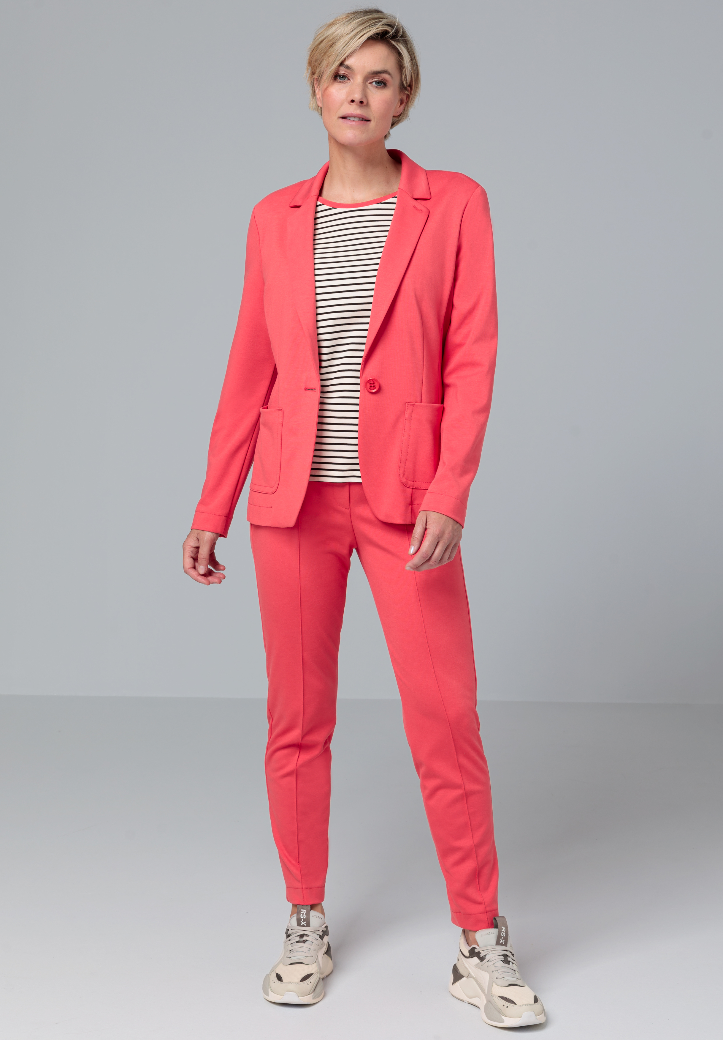 Eine Person mit kurzen blonden Haaren trägt einen korallenrosa Anzug mit passendem Blazer und Hose, ein schwarz-weiß gestreiftes Hemd und beige Turnschuhe und steht vor einem schlichten hellgrauen Hintergrund.