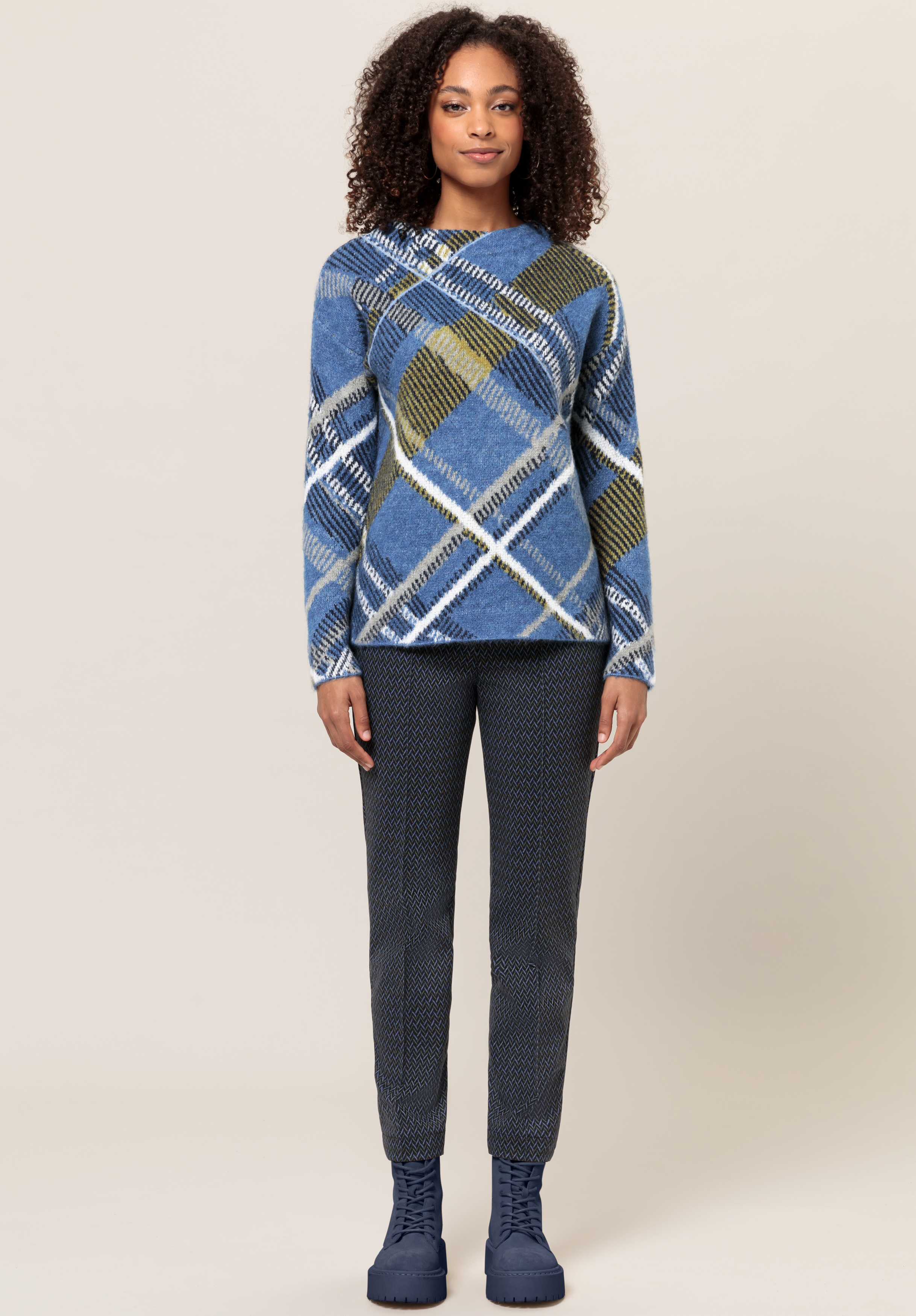 Eine Frau steht mit Blick nach vorne und trägt einen blau-grün-weiß-grauen karierten Pullover, eine dunkel gemusterte Hose und blaue Schnürstiefel vor einem schlichten beigen Hintergrund.