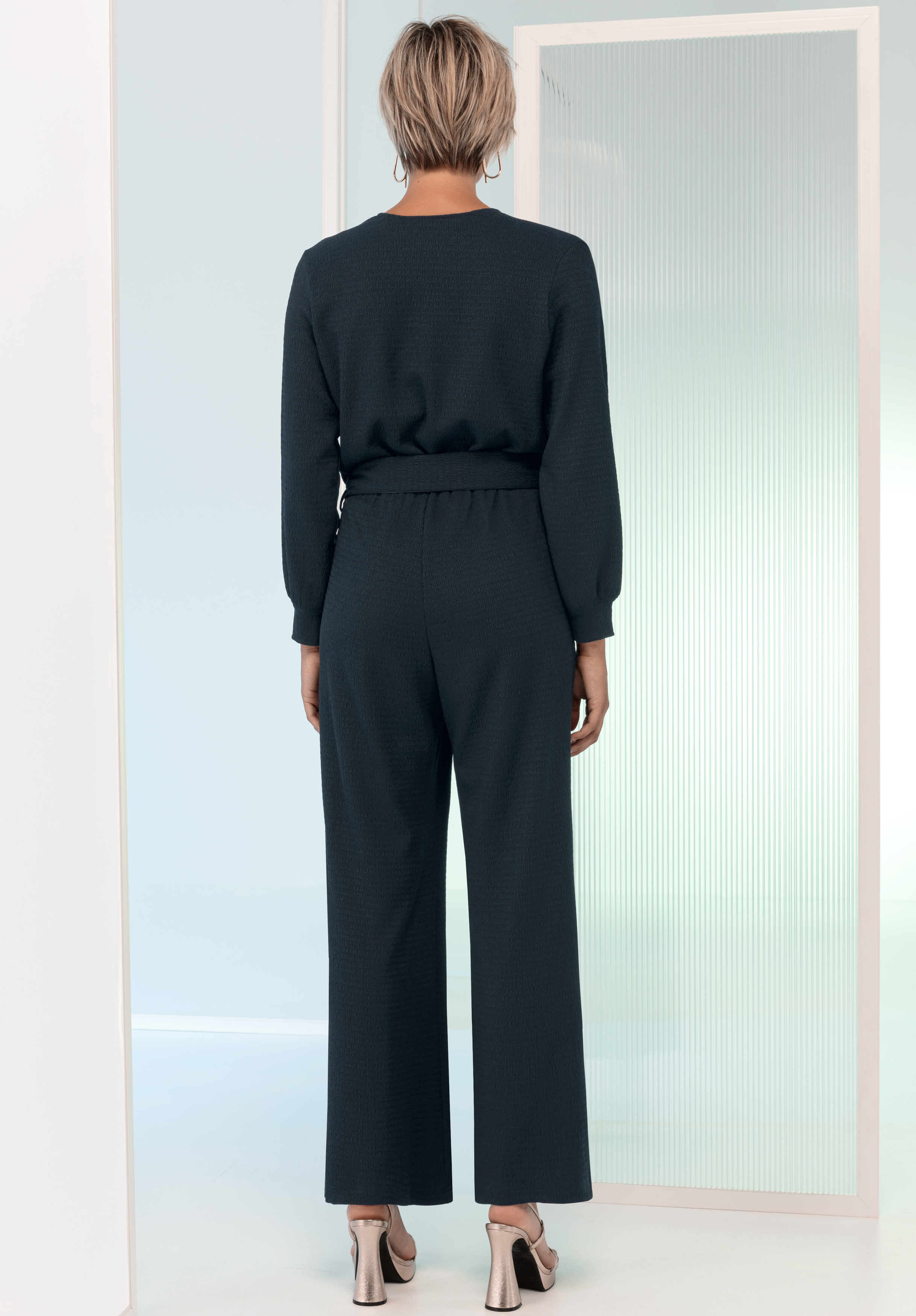 Eine Person mit kurzen, hellen Haaren steht mit dem Rücken zur Kamera, trägt einen langärmeligen, dunklen Overall und Sandalen mit Absatz in einem minimalistisch-modernen Raum mit hellen Wänden und einer Milchglasscheibe.