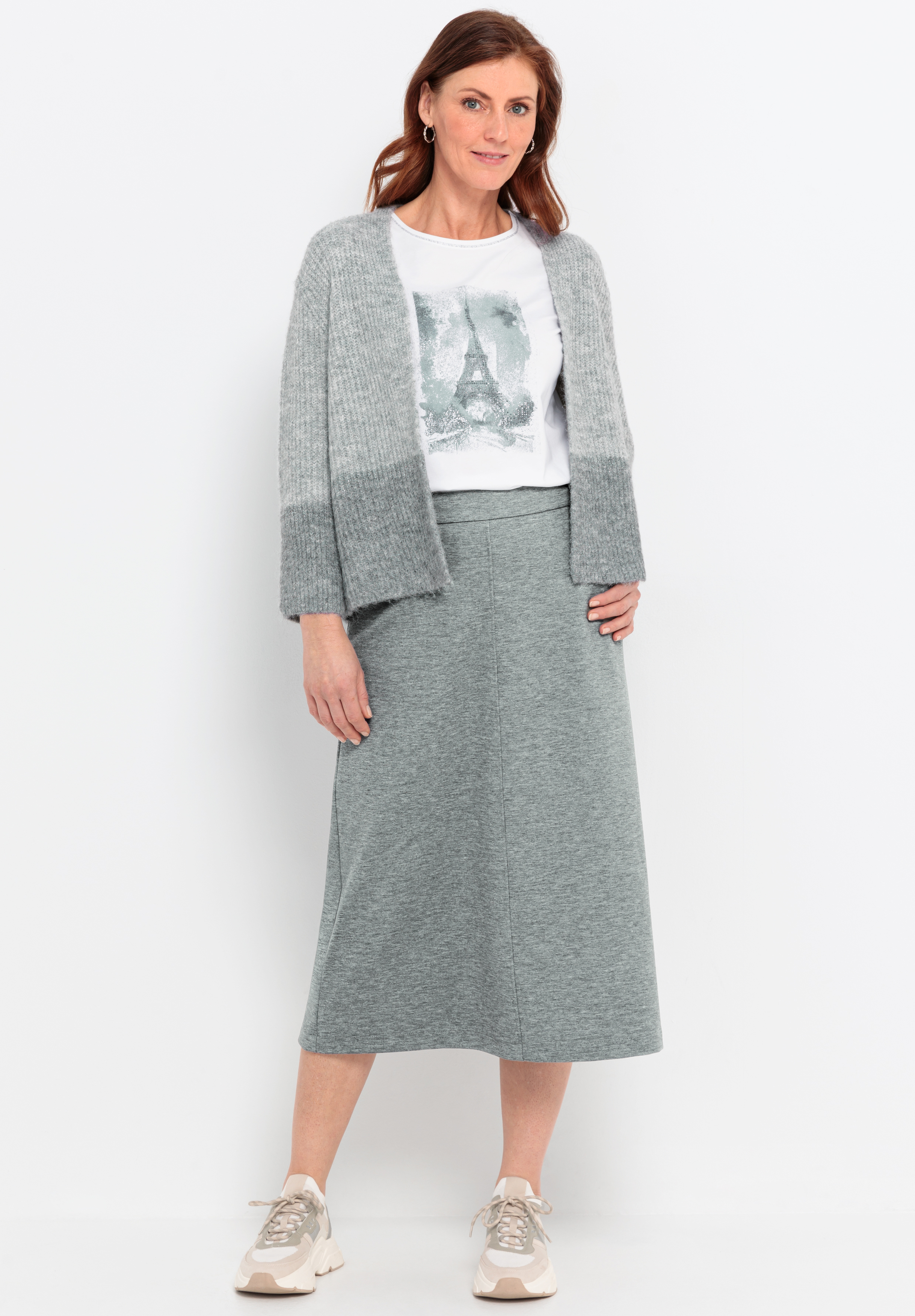 Eine Frau steht vor einem weißen Hintergrund und trägt eine graue Strickjacke, ein weißes T-Shirt mit einer Eiffelturm-Grafik, einen grauen Midi-Rock und beigefarbene Turnschuhe. Sie hat schulterlanges kastanienbraunes Haar und lächelt leicht.