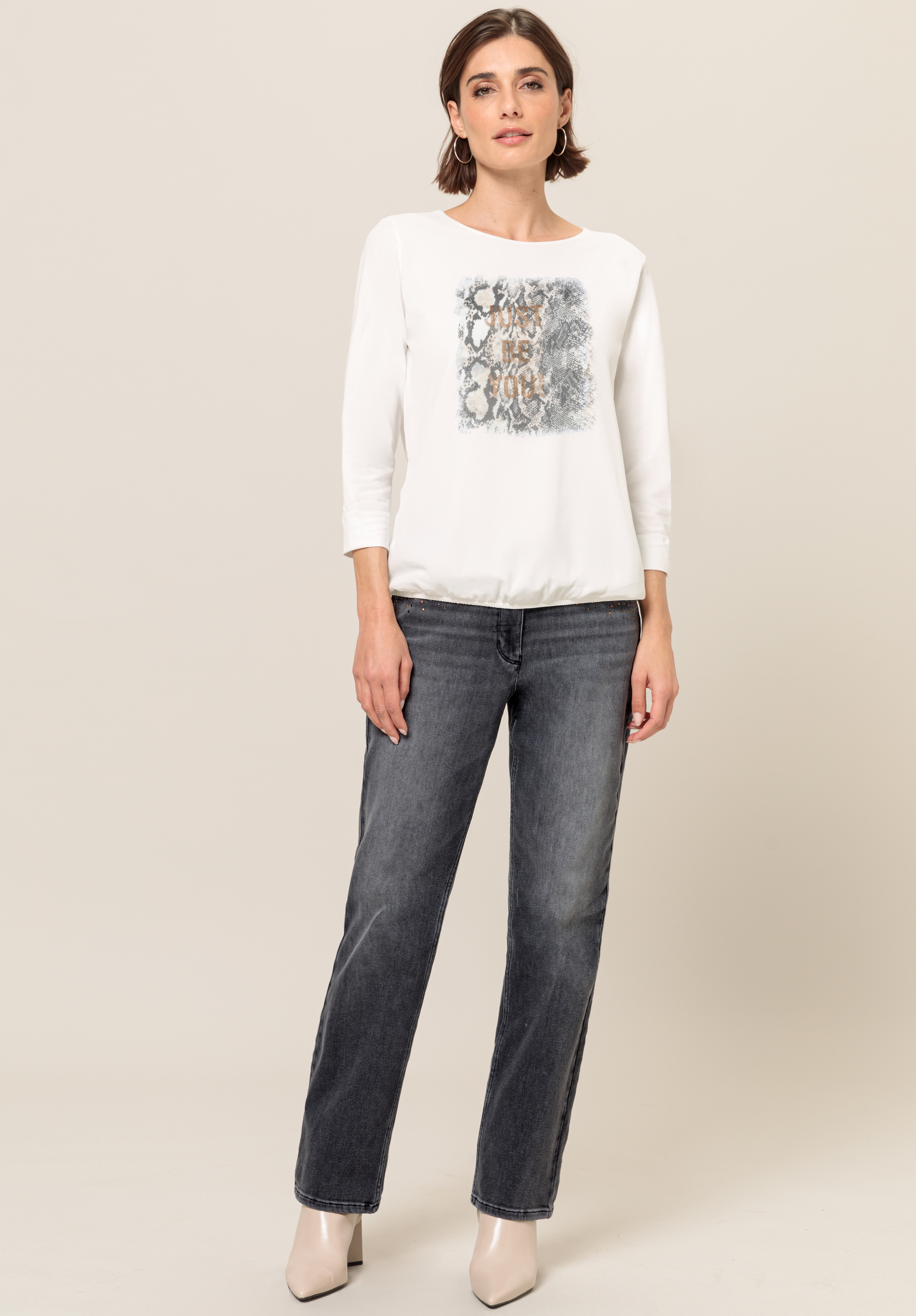 Vor einem schlichten Hintergrund steht eine Frau, die ein weißes Langarmshirt mit einer rechteckigen abstrakten Grafik, dunkle Jeans mit geradem Bein und helle Schuhe mit Absatz trägt.