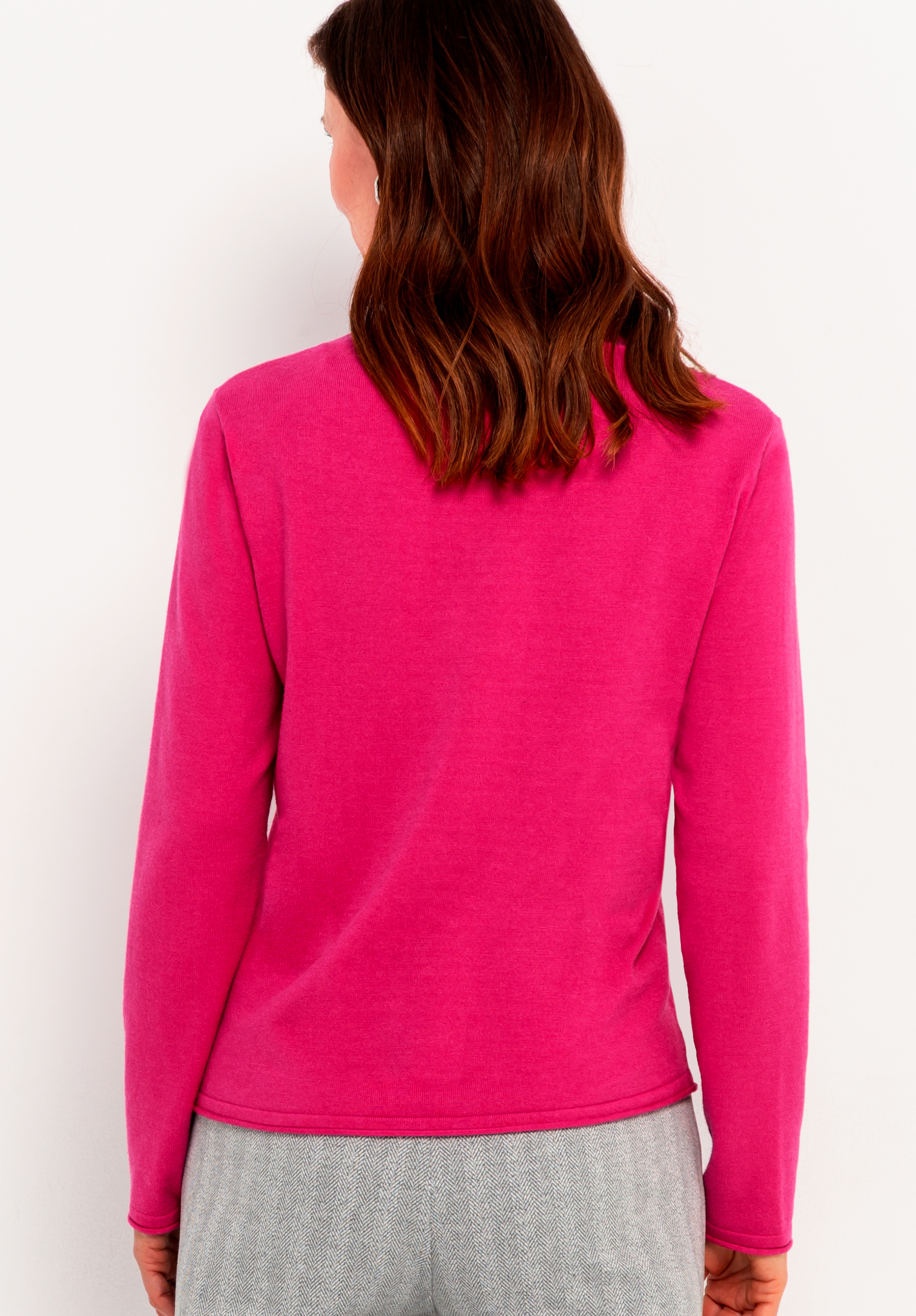 Eine Frau mit mittellangem braunem Haar ist von hinten zu sehen. Sie trägt ein langärmeliges, hellrosa Oberteil und eine hellgraue Hose vor einem schlichten weißen Hintergrund.