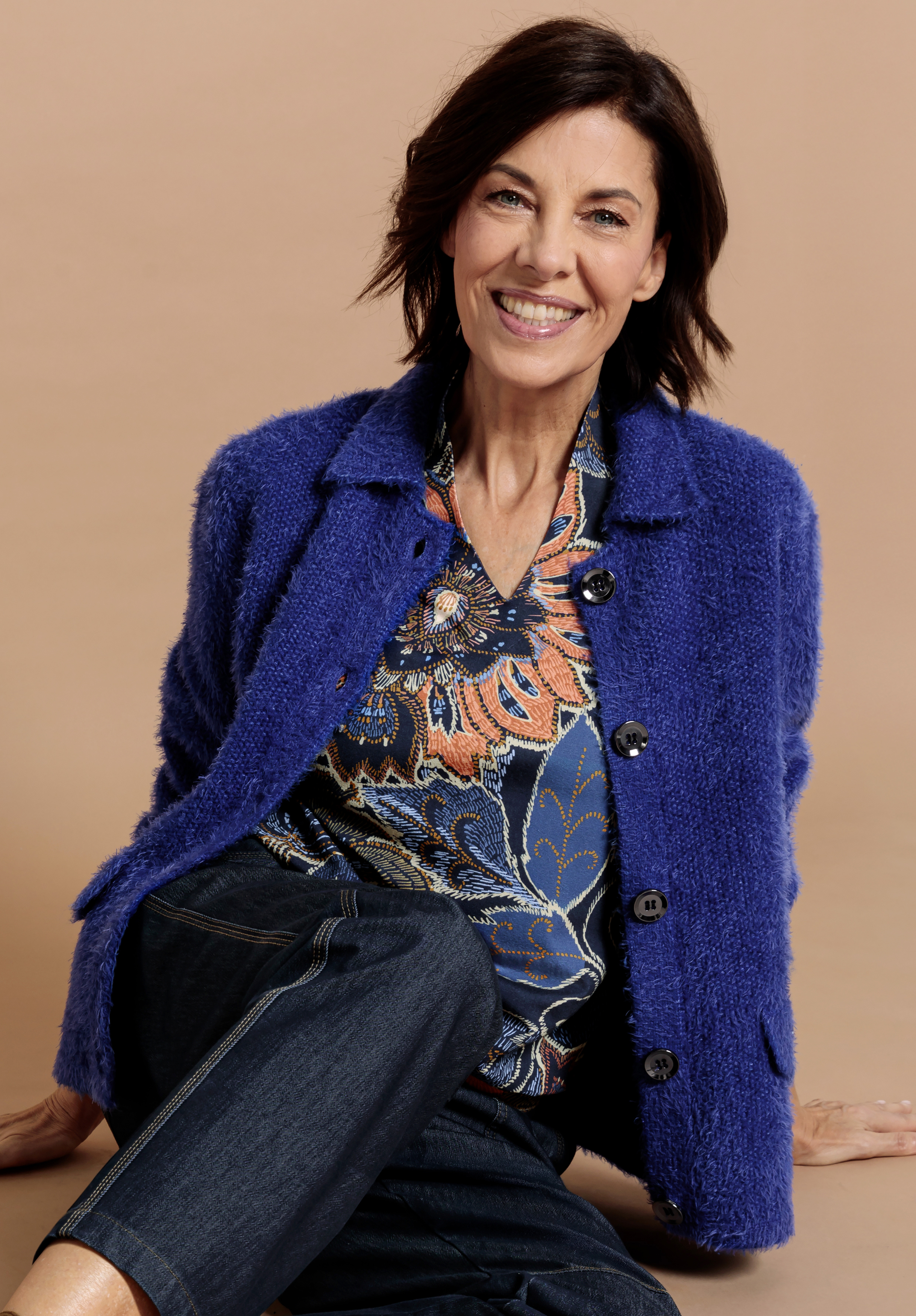 Eine lächelnde Frau mit kurzen dunklen Haaren trägt eine blaue strukturierte Jacke über einer geblümten Bluse und dunkle Jeans und sitzt lässig vor einem beigen Hintergrund.