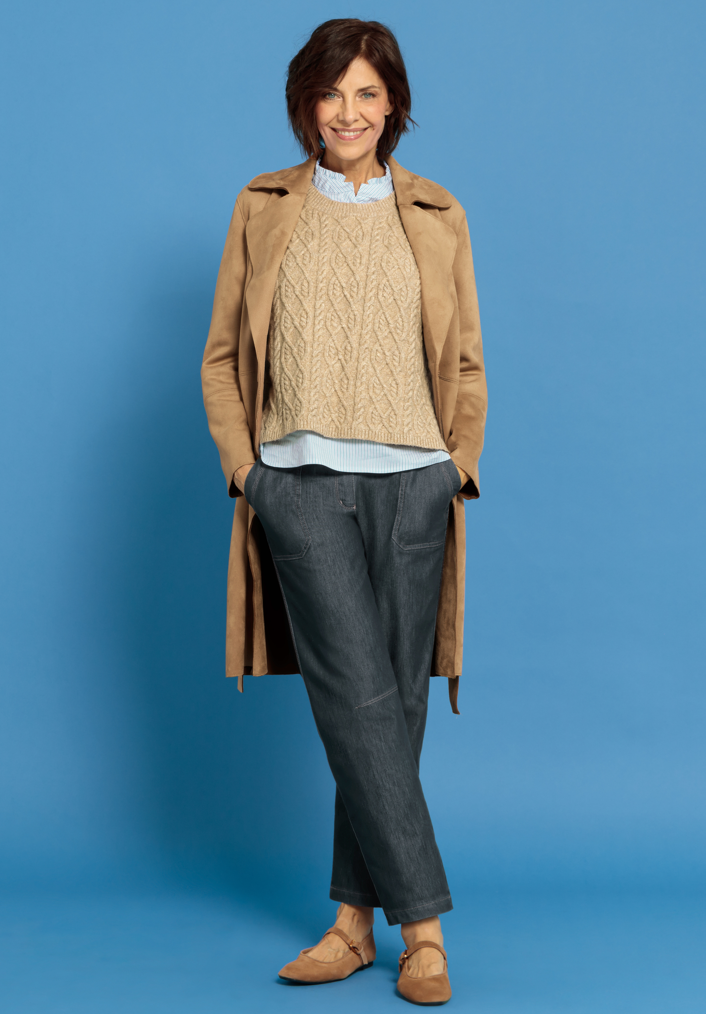 Eine Frau steht vor einem blauen Hintergrund, trägt einen hellbraunen Mantel, einen beigen Zopfstrickpullover über einem hellblauen Hemd, dunkle Jeans und hellbraune Slipper und lächelt mit den Händen in den Taschen.