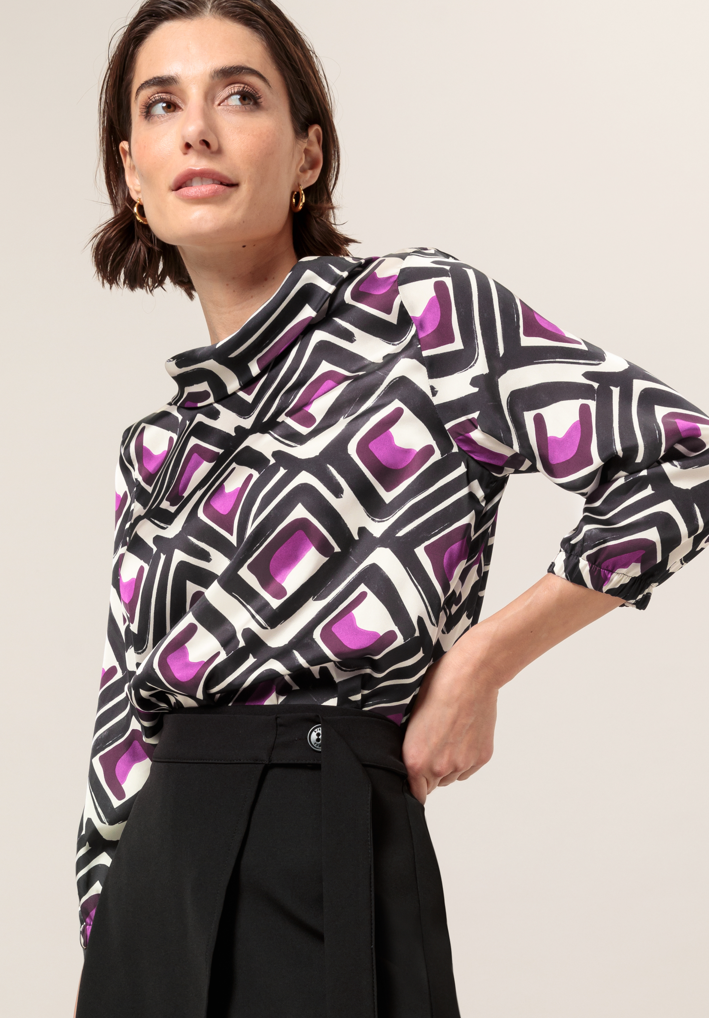 Eine Frau mit kurzen braunen Haaren trägt eine gemusterte Bluse mit geometrischen Formen in Schwarz, Weiß und Lila und dazu eine hoch taillierte schwarze Hose.