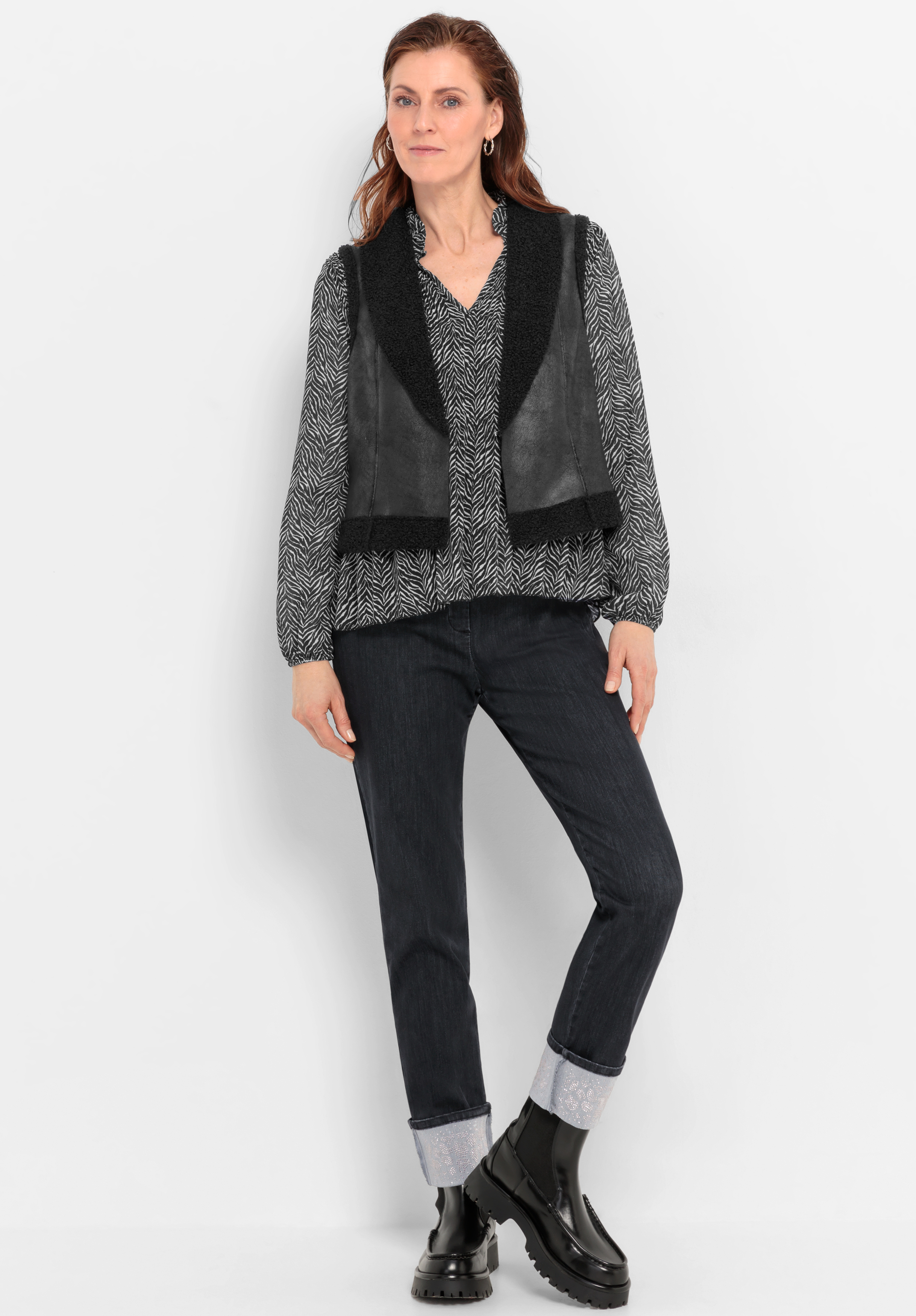 Vor einem weißen Hintergrund steht eine Frau, die eine schwarz-weiß gemusterte Bluse, eine schwarze Weste, eine dunkle Jeans mit Umschlag und schwarze Stiefel trägt. Sie hat langes braunes Haar und eine entspannte, selbstbewusste Haltung.