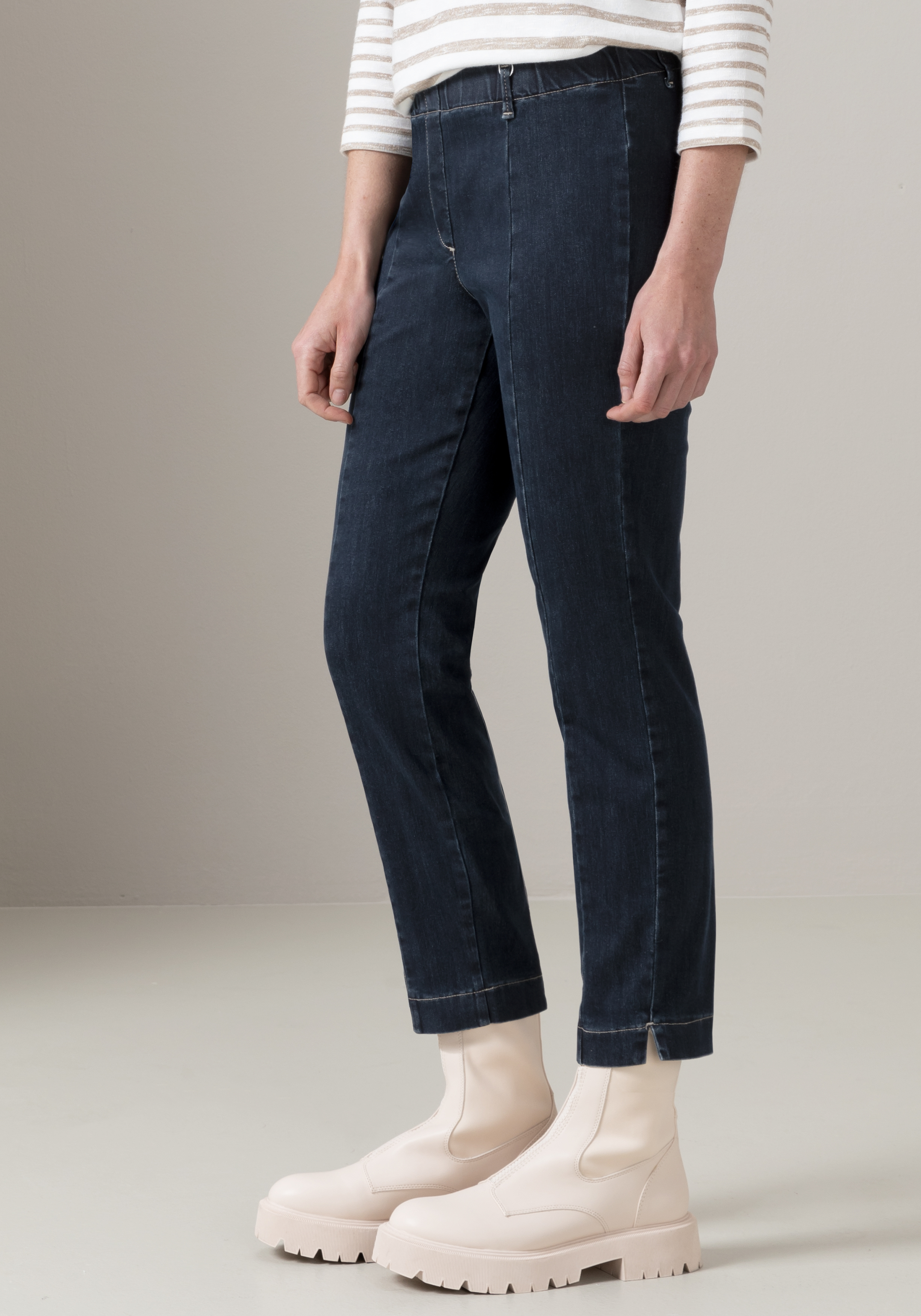 Eine Person, die dunkelblaue Jeans, ein weiß-beige gestreiftes Langarmshirt und klobige, cremefarbene Stiefel trägt, steht vor einem schlichten, hellen Hintergrund.