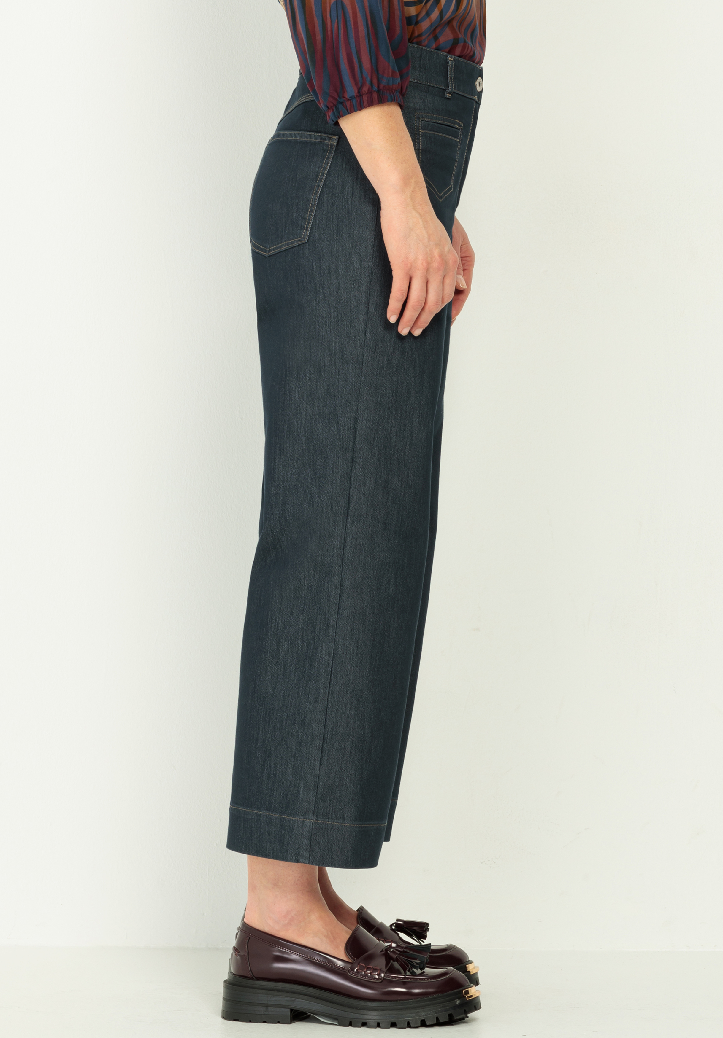 Eine seitlich stehende Person trägt dunkelblaue Jeans mit geradem Bein, ein gestreiftes langärmeliges Hemd und weinrote Halbschuhe mit dekorativen Details. Die Umgebung ist ein schlichter, heller Hintergrund.