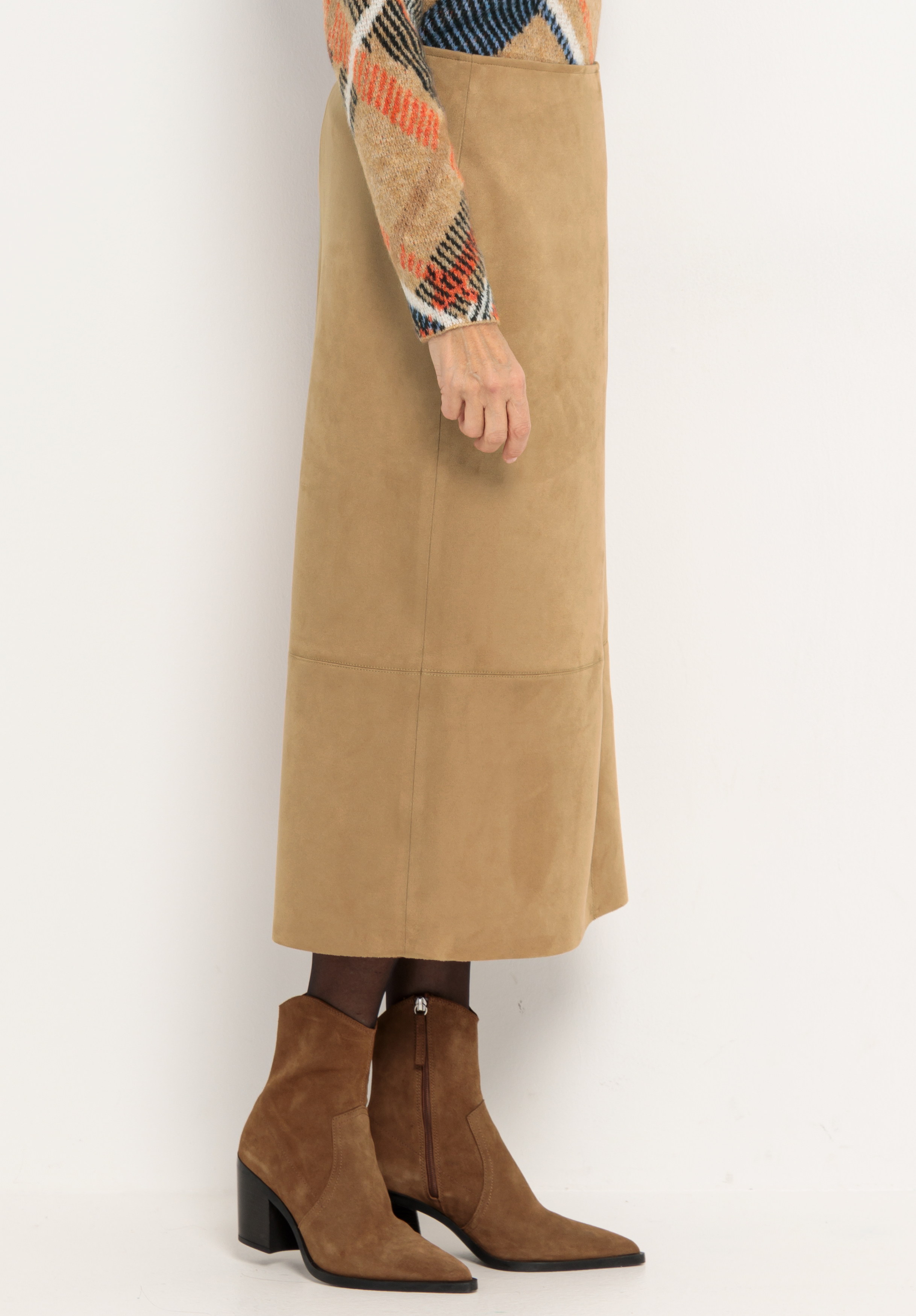 Eine Person, die einen hellbraunen Midirock aus Wildleder, braune spitze Wildlederstiefel mit Blockabsatz und einen bunt gemusterten Pullover trägt, steht vor einem schlichten hellen Hintergrund.