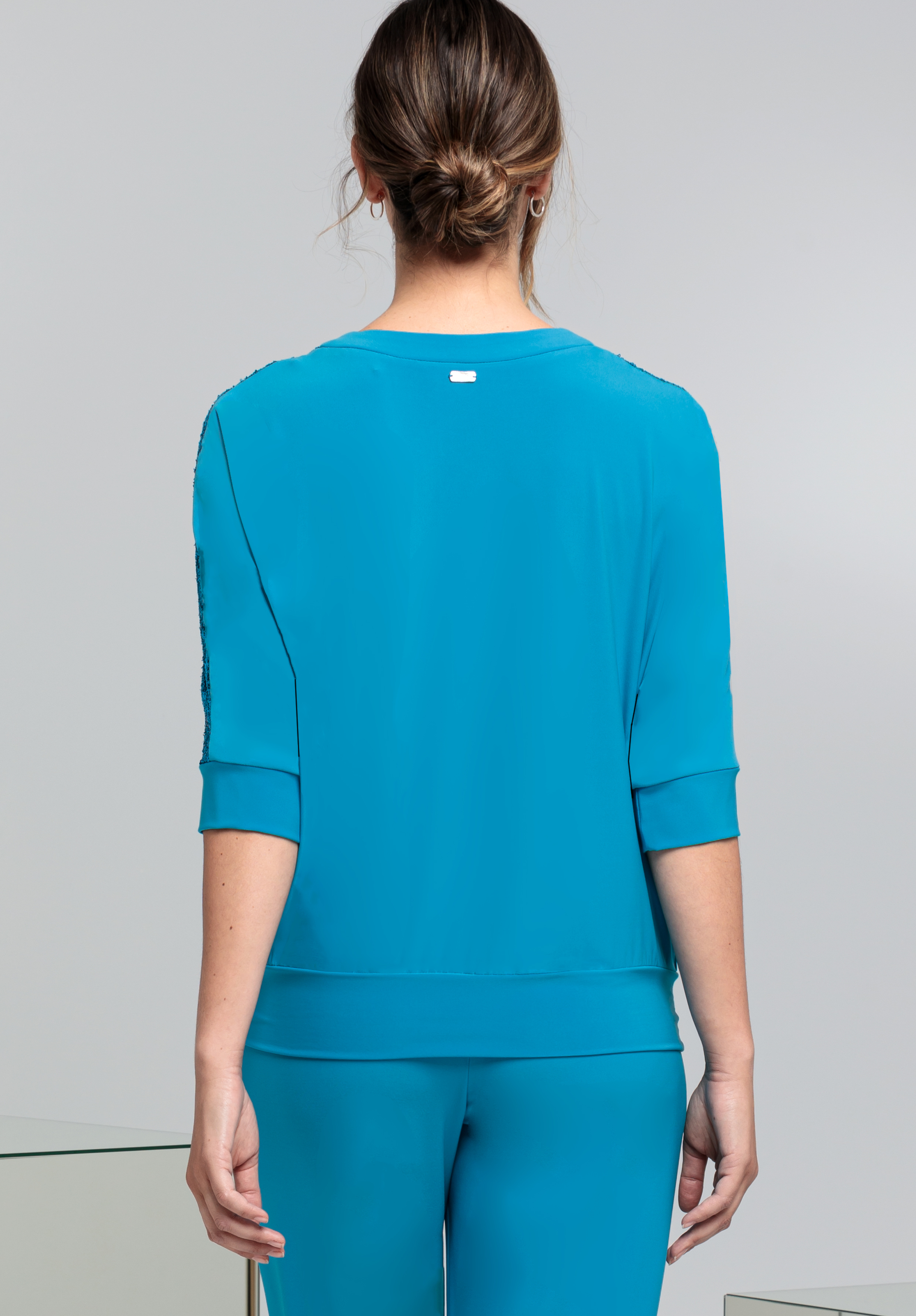 Eine Frau mit braunem Haar, tief in einem gedrehten Dutt, steht mit dem Rücken zur Kamera. Sie trägt ein leuchtend blaues Oberteil mit Dreiviertelärmeln und eine dazu passende Hose. Der Hintergrund ist schlicht und hellgrau.
