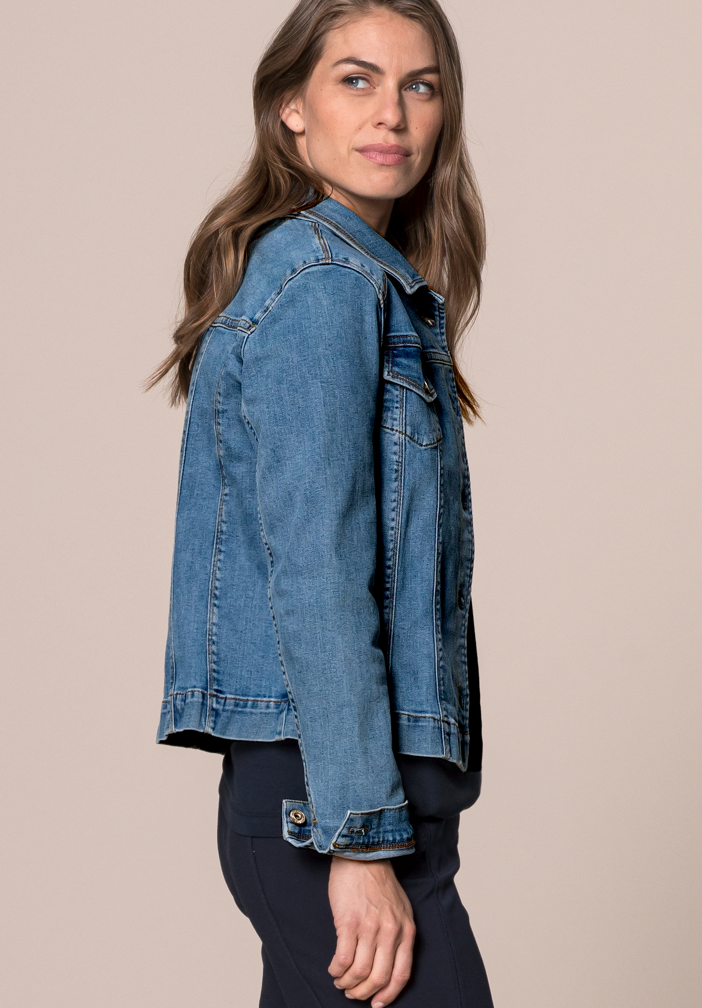 Eine Frau mit langen braunen Haaren, die eine blaue Jeansjacke und eine dunkle Hose trägt, steht mit dem Gesicht nach links, blickt über die Schulter und lächelt leicht vor einem schlichten beigen Hintergrund.