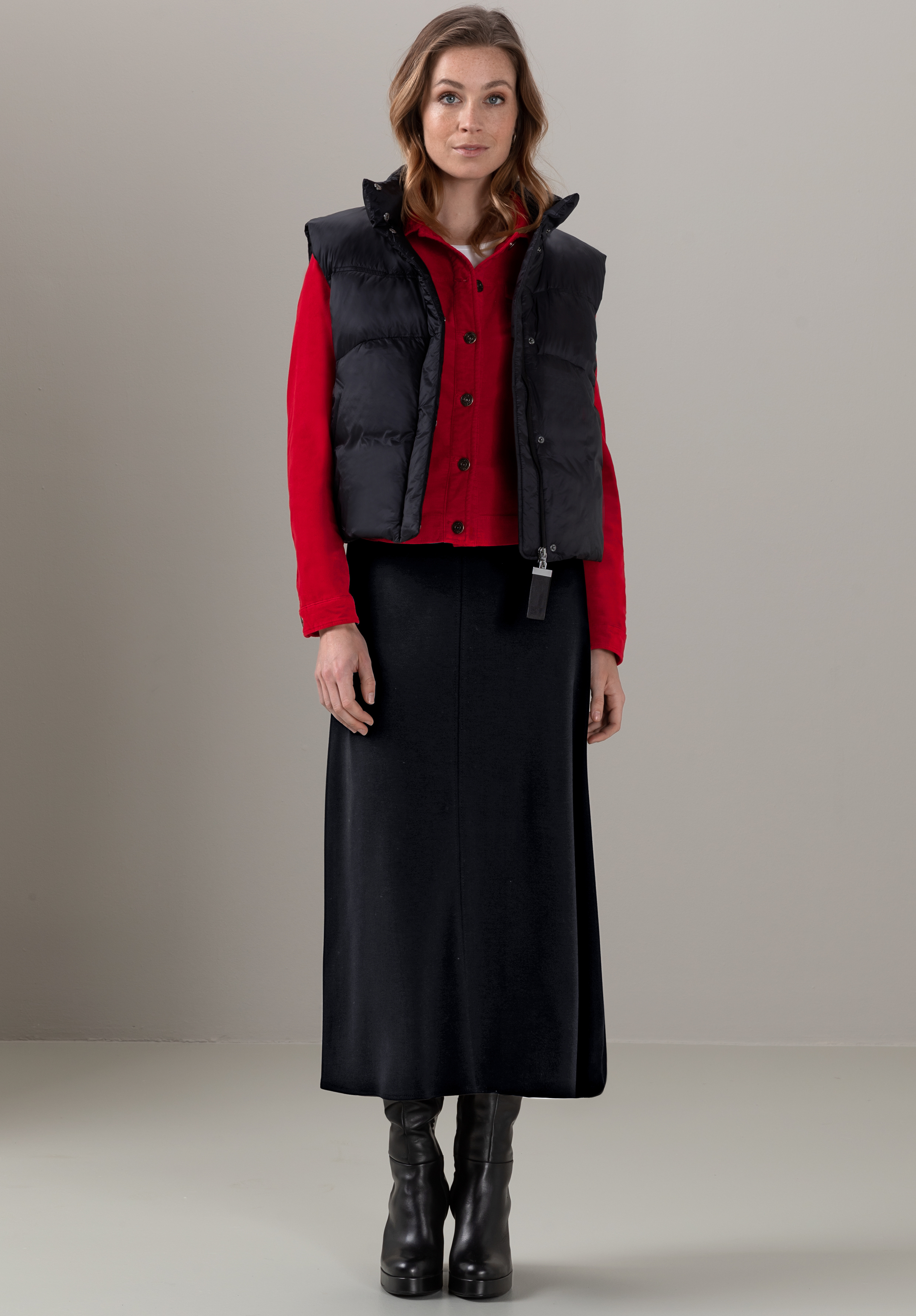 Eine Frau steht mit dem Gesicht nach vorne. Sie trägt eine schwarze Steppweste über einem roten Hemd, einen langen schwarzen Rock und schwarze Stiefel mit Absatz. Sie hat hellbraunes Haar und einen neutralen Gesichtsausdruck.