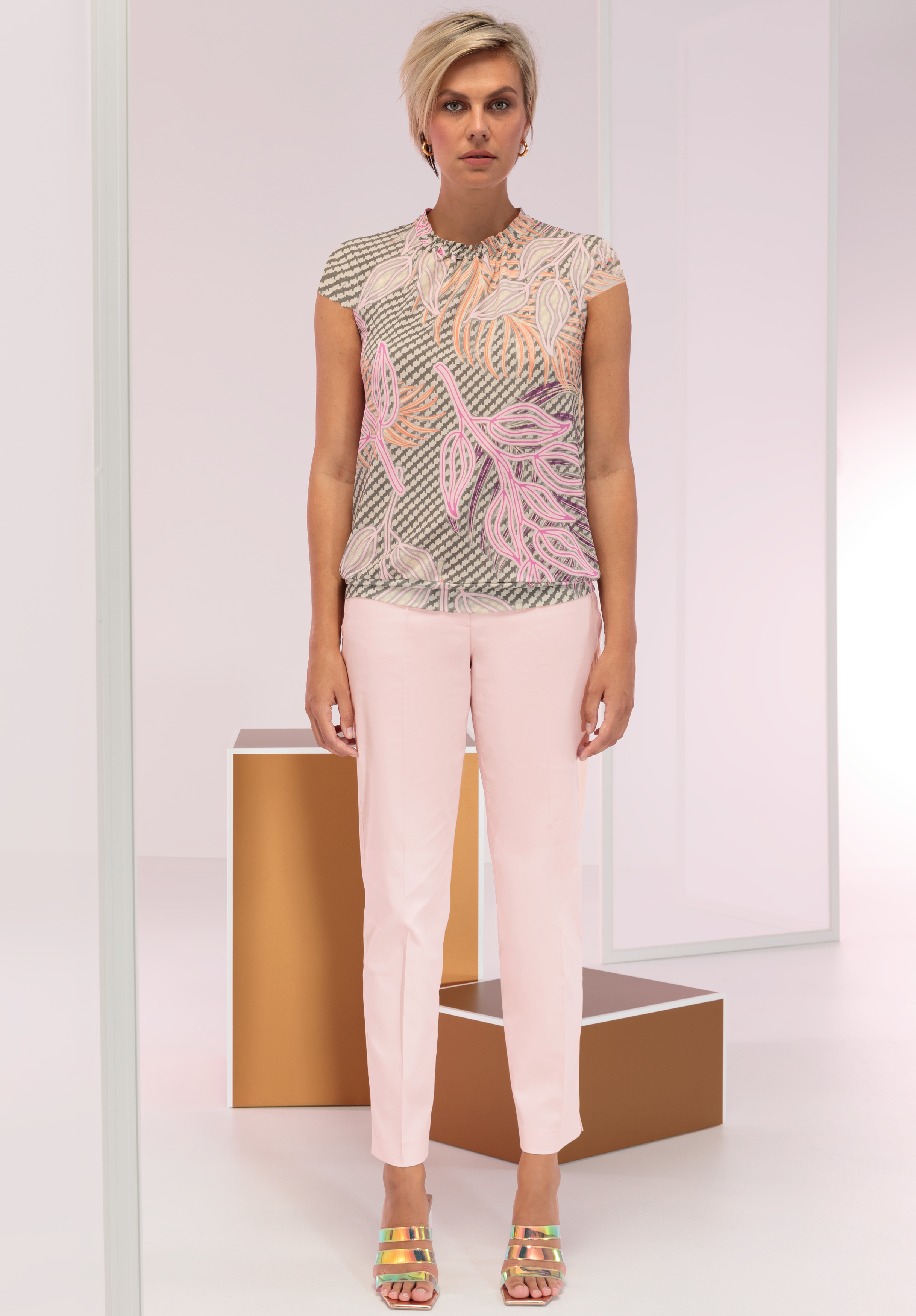 Eine Frau mit kurzen blonden Haaren trägt ein gemustertes Oberteil in Rosa- und Beigetönen, eine hellrosa Hose und bunte Sandalen. Sie steht in einer minimalistischen Umgebung mit geometrischen Formen in Gold und Weiß im Hintergrund.