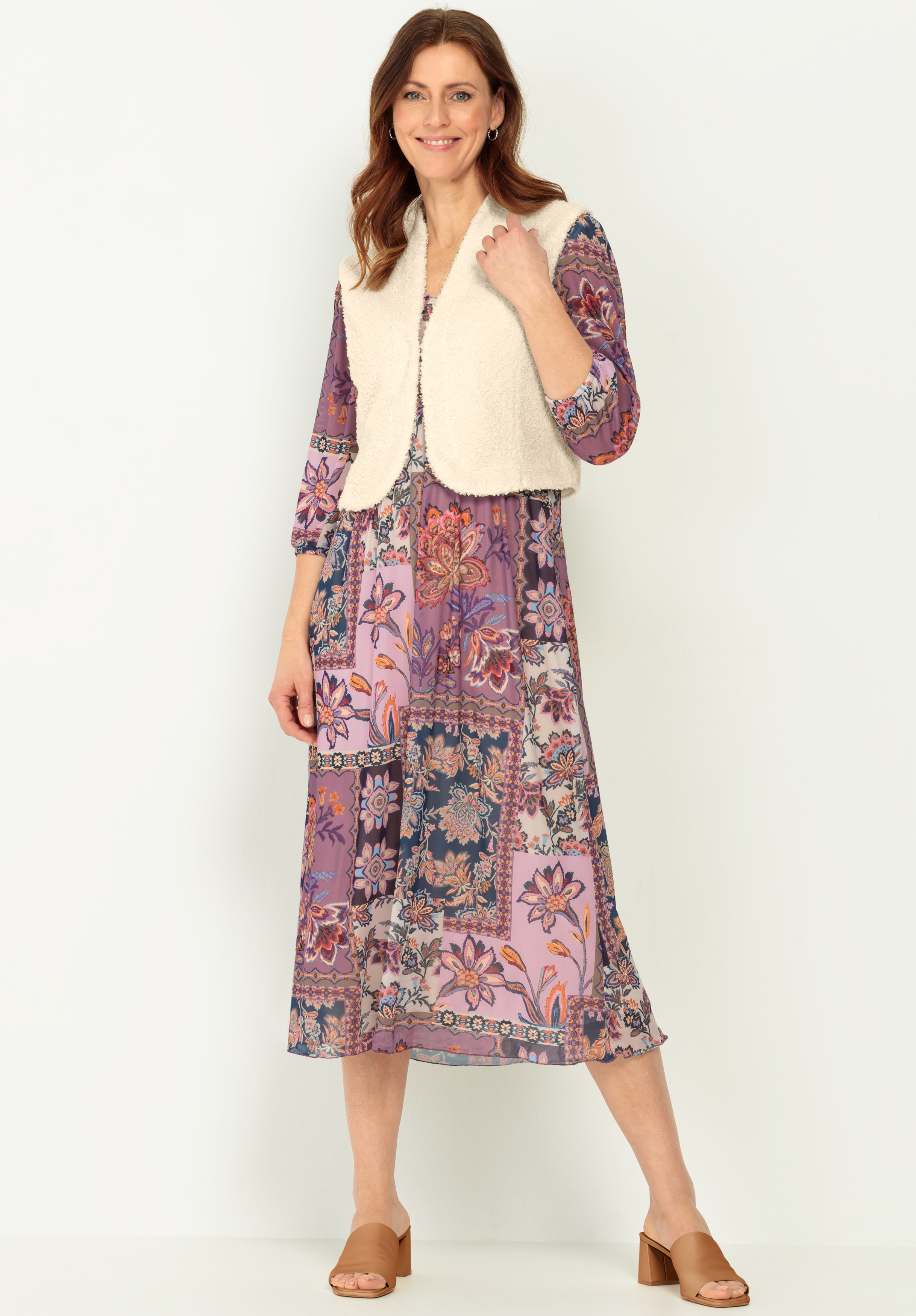 Eine Frau, die eine cremefarbene, wattierte Weste über einem langen, lilafarbenen, geblümten Patchwork-Kleid und hellbraune, hochhackige Sandalen trägt, steht lächelnd vor einem schlichten, hellen Hintergrund.