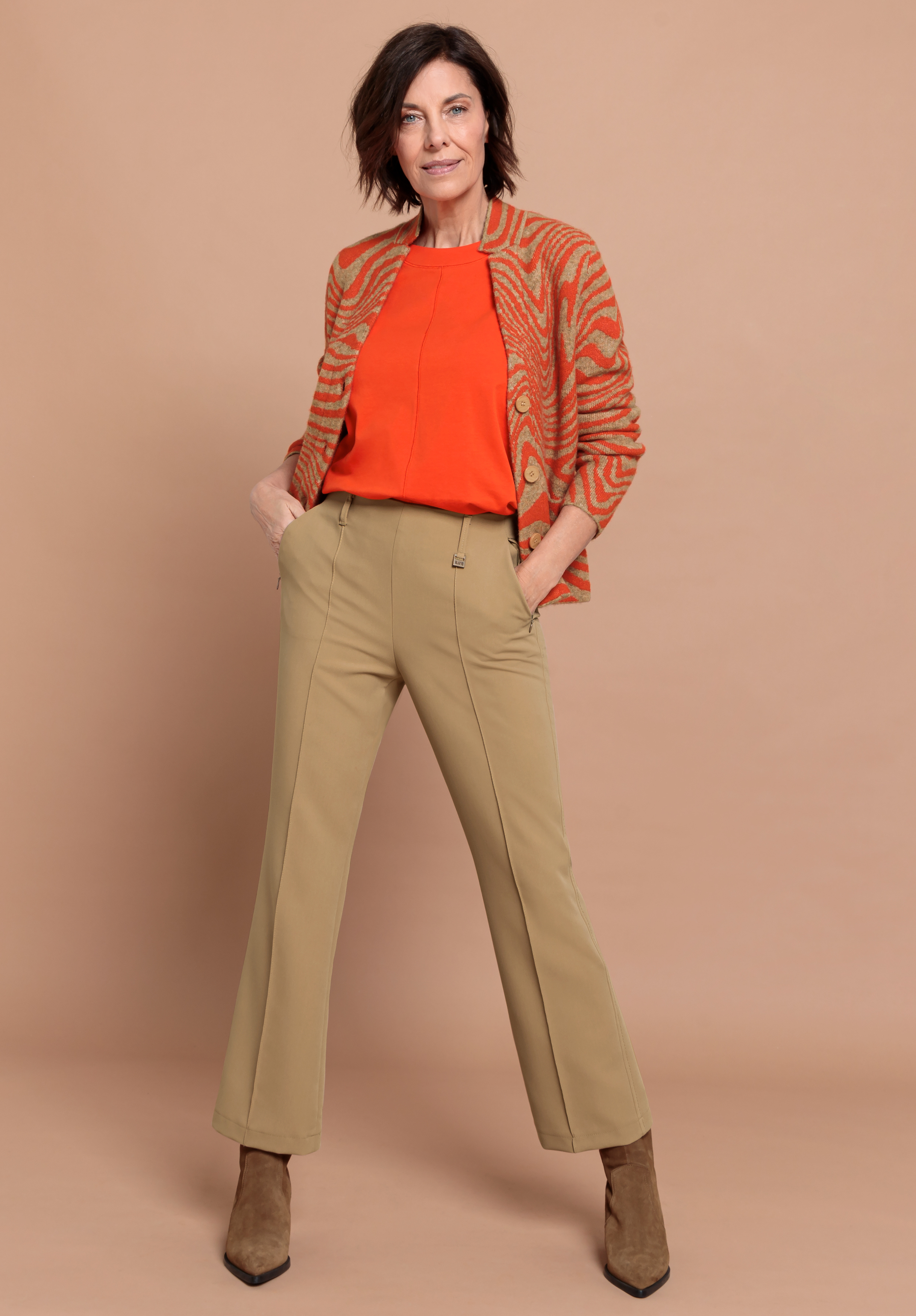 Eine Frau steht vor einem beigen Hintergrund, trägt eine rost- und beigegemusterte Strickjacke, ein orangefarbenes Oberteil, eine olivgrüne Hose und hellbraune Stiefeletten, hat die Hände in den Taschen und schaut selbstbewusst in die Kamera.