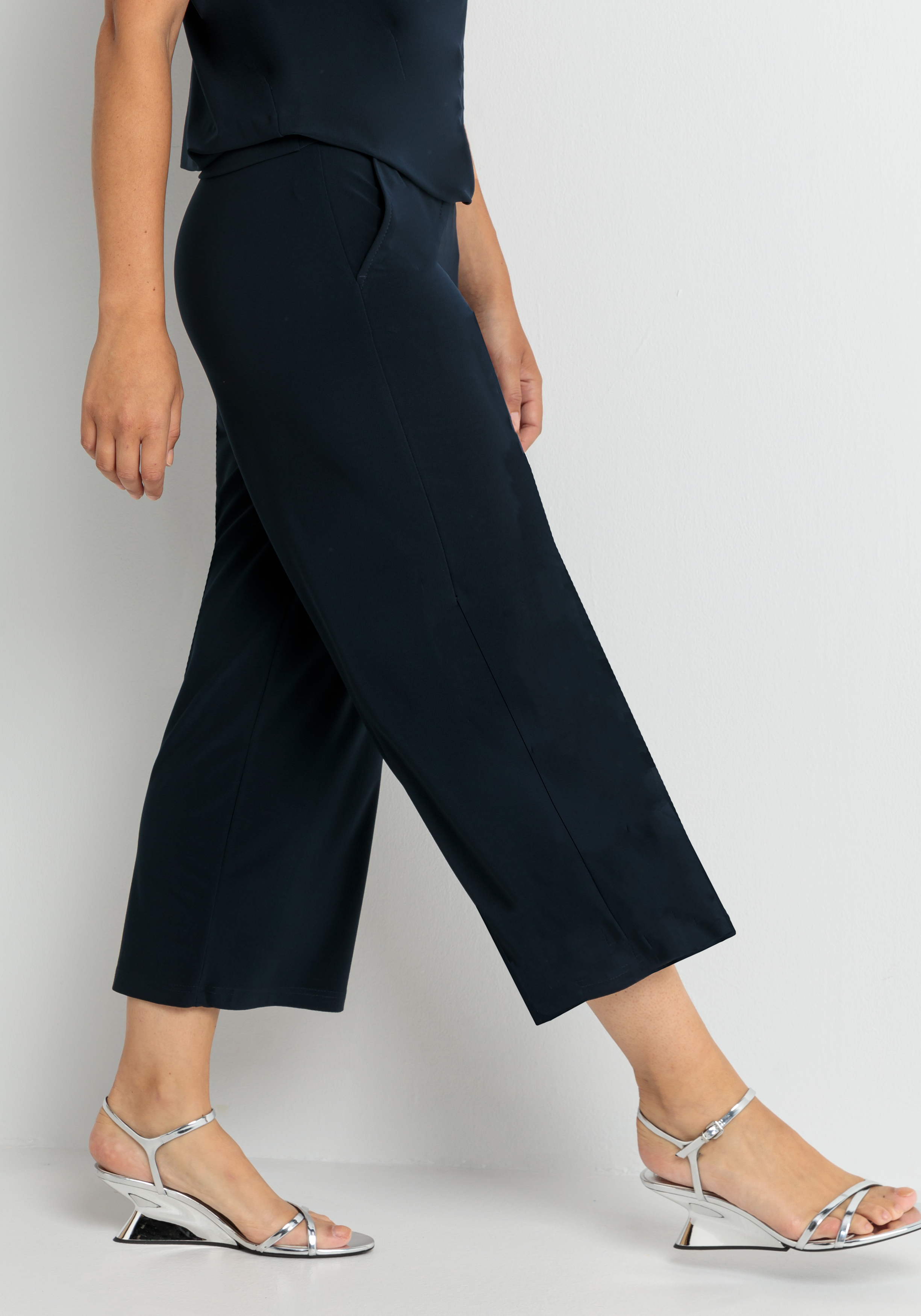 Eine Person, die eine marineblaue Hose mit weitem Bein, ein passendes Oberteil und silberne Riemchensandalen mit niedrigen Absätzen trägt, wird vor einem hellen Hintergrund gezeigt.