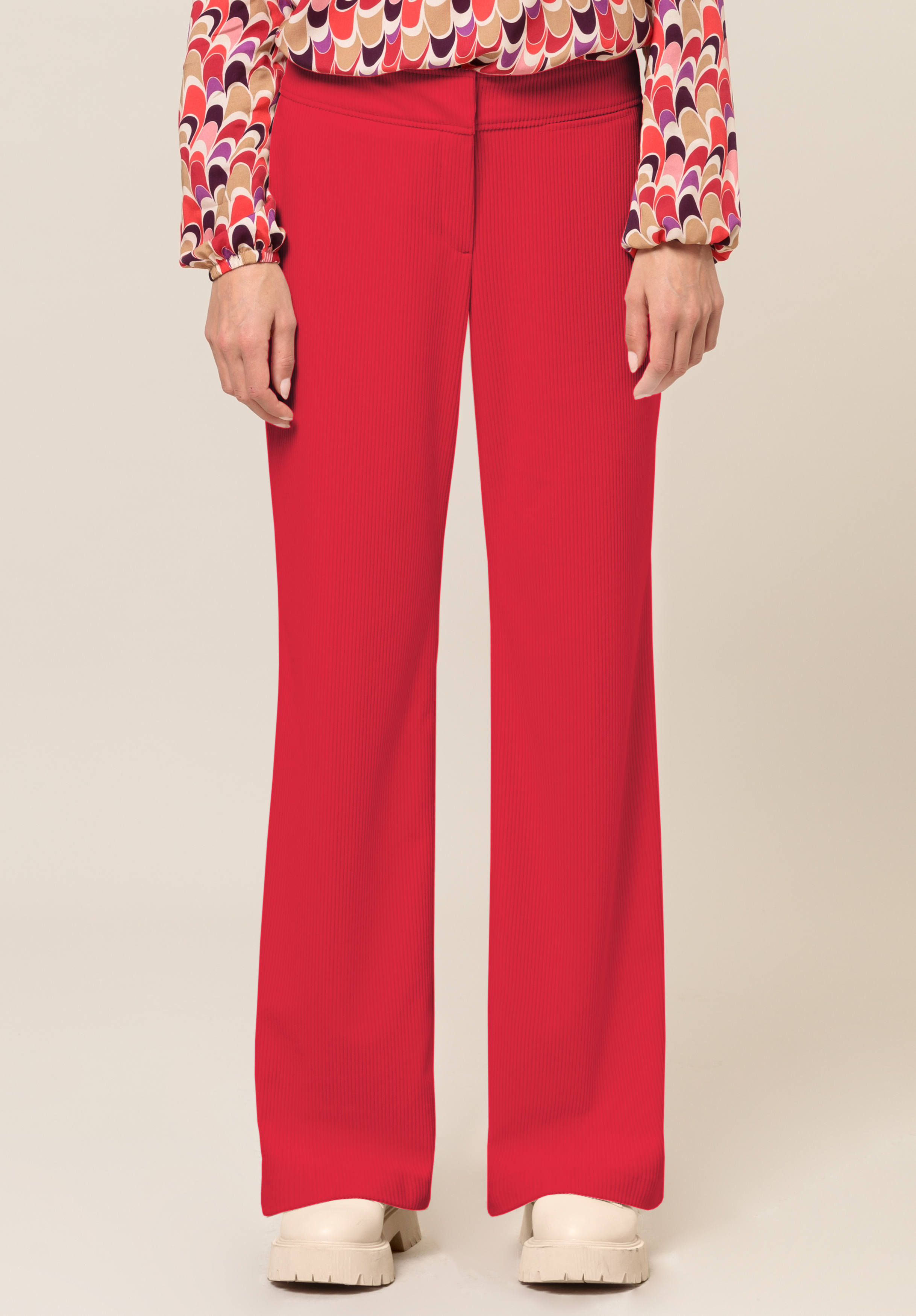 Cordhose PARIGI Eine Person trägt leuchtend rote Hosen mit weitem Bein, ein buntes, langärmliges, gemustertes Oberteil und cremefarbene, klobige Schuhe und steht vor einem schlichten, hellen Hintergrund.