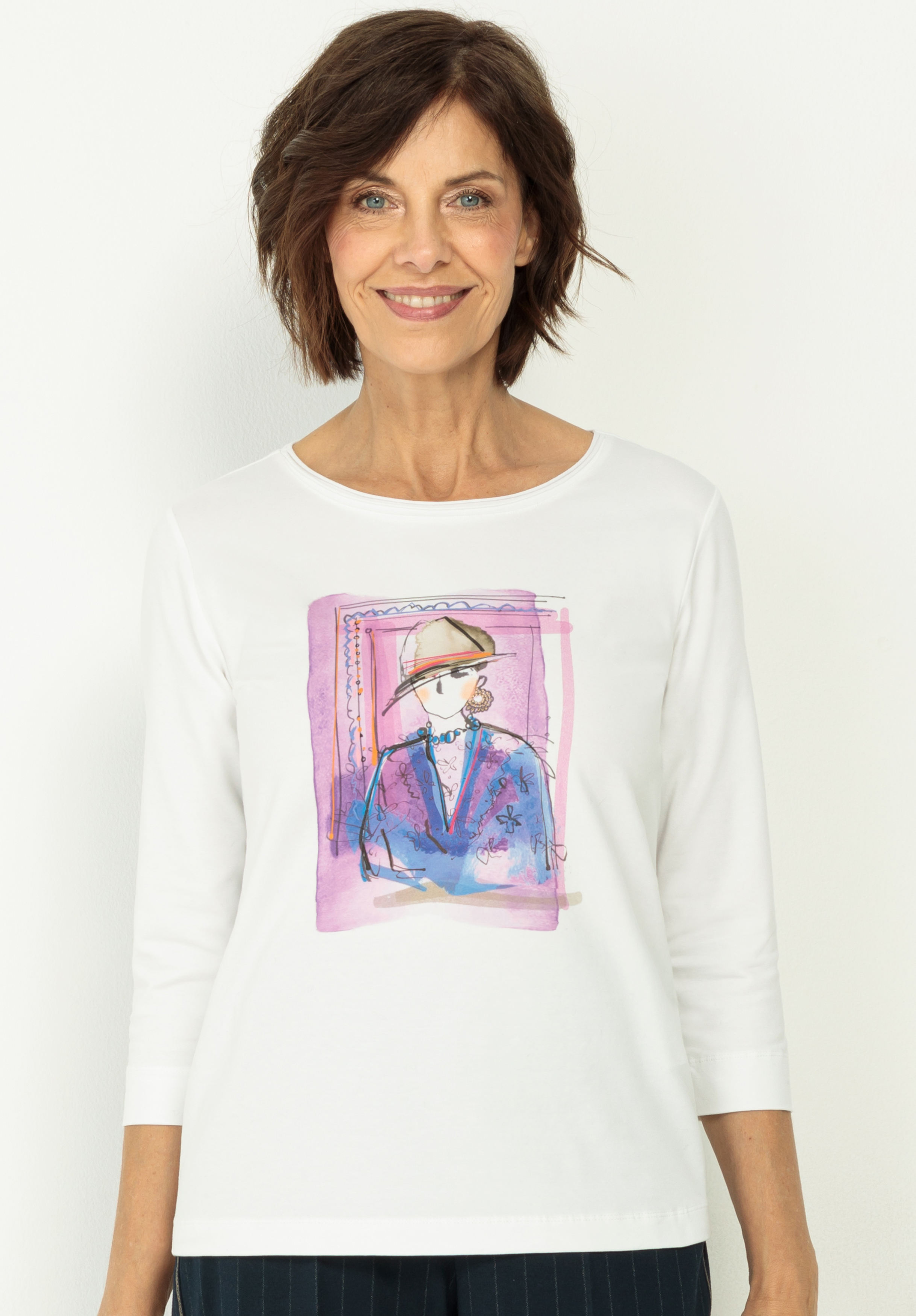 Rundhalsshirt DINI Eine Frau mit kurzen braunen Haaren lächelt und trägt ein weißes langärmeliges Hemd, auf dessen Vorderseite eine bunte Skizze einer Person mit blauem Mantel und Hut abgebildet ist.