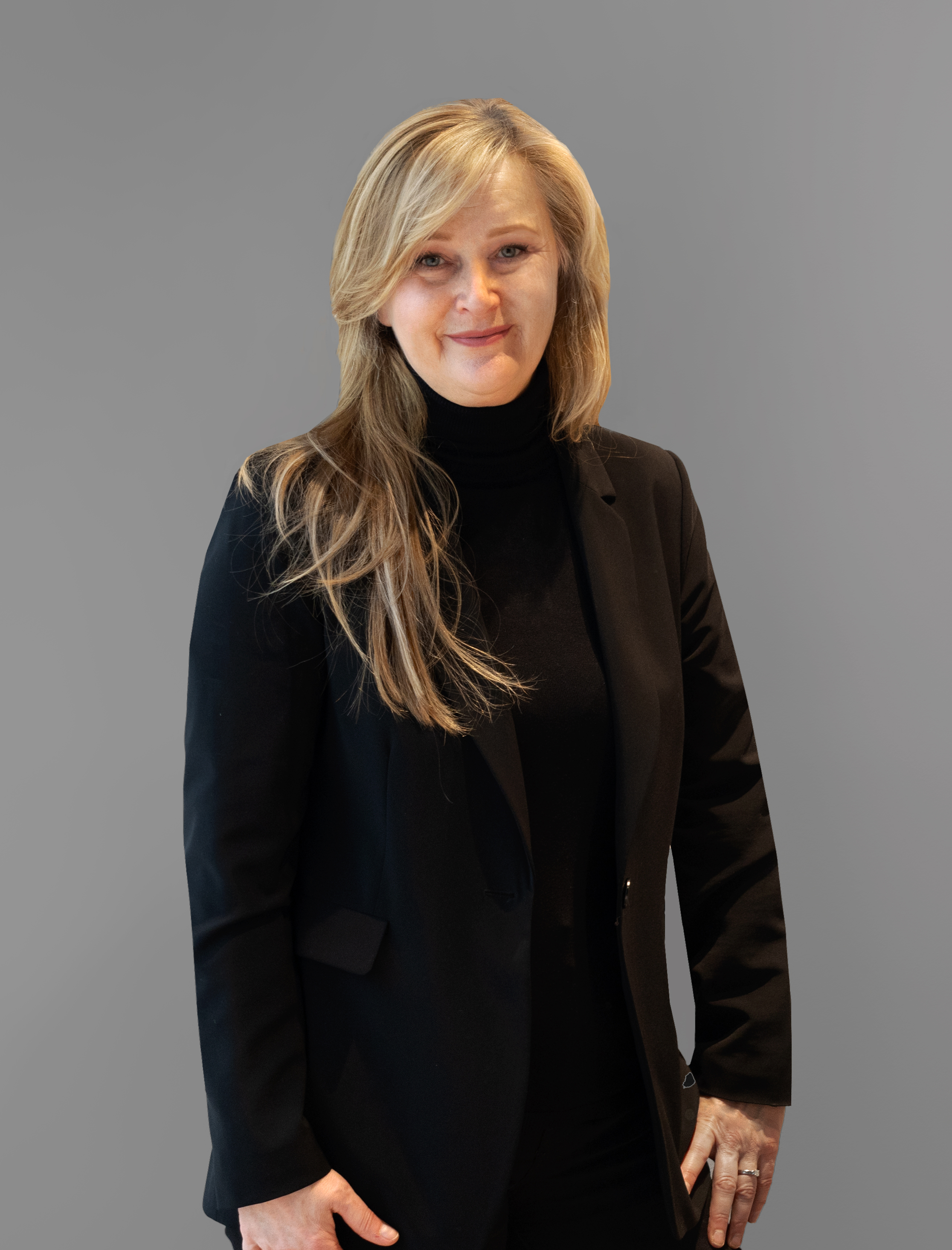 Eine Frau mit langen blonden Haaren, einem schwarzen Blazer und einem schwarzen Rollkragenpullover steht vor einem schlichten grauen Hintergrund und lächelt leicht, während ihre Hände an den Seiten hängen.