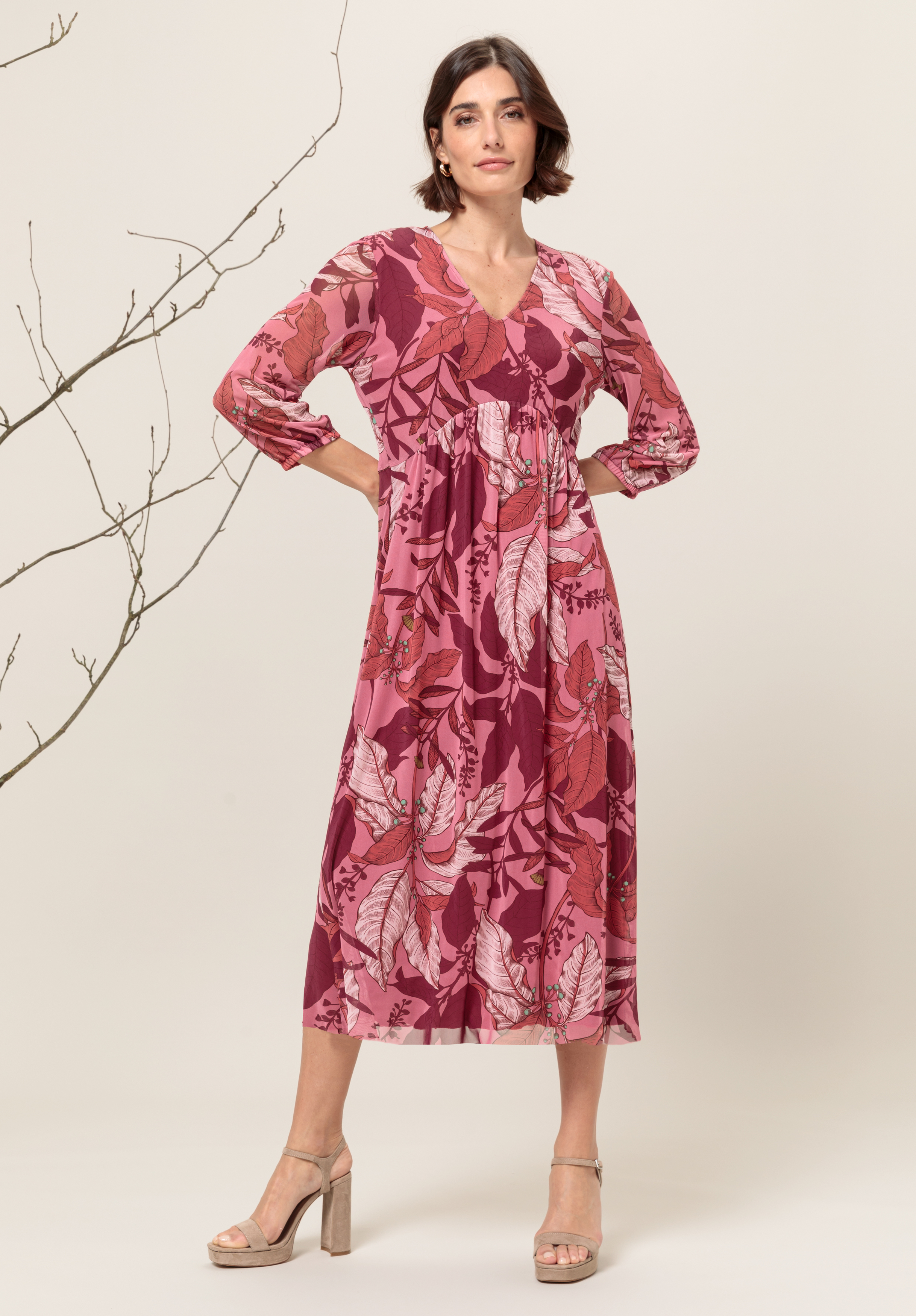Eine Frau steht selbstbewusst in einem fließenden, langärmeligen, rosa-roten Blumenkleid mit V-Ausschnitt und Midi-Länge, kombiniert mit beigen High Heels. Sie posiert vor einem neutralen Hintergrund mit kahlen Ästen.
