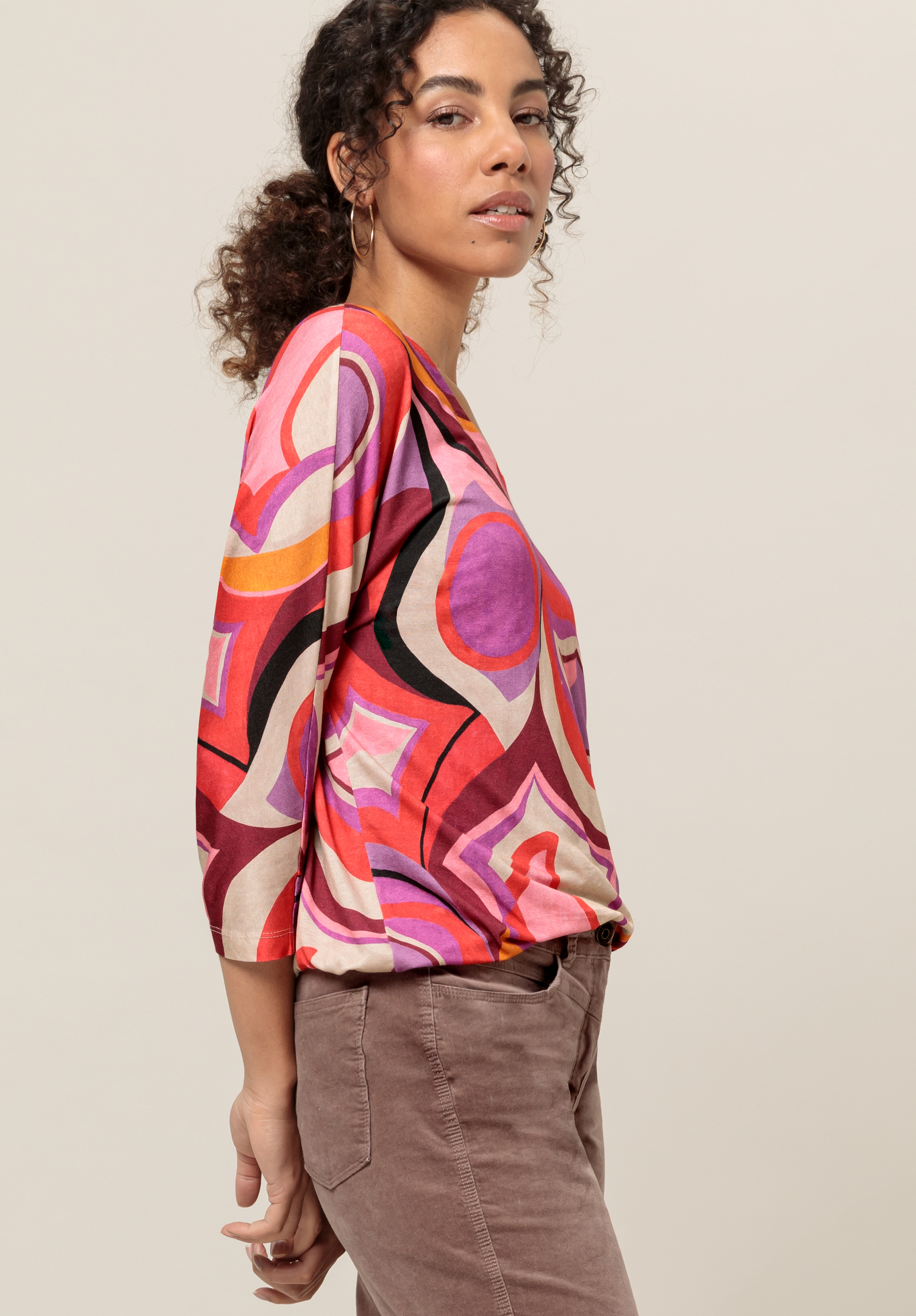 Eine Frau mit lockigem Haar trägt ein farbenfrohes Oberteil mit geometrischem Muster in Rot-, Rosa- und Lilatönen, dazu eine hoch taillierte, hellbraune Hose. Sie steht seitlich und blickt selbstbewusst vor einem schlichten Hintergrund in die Kamera.