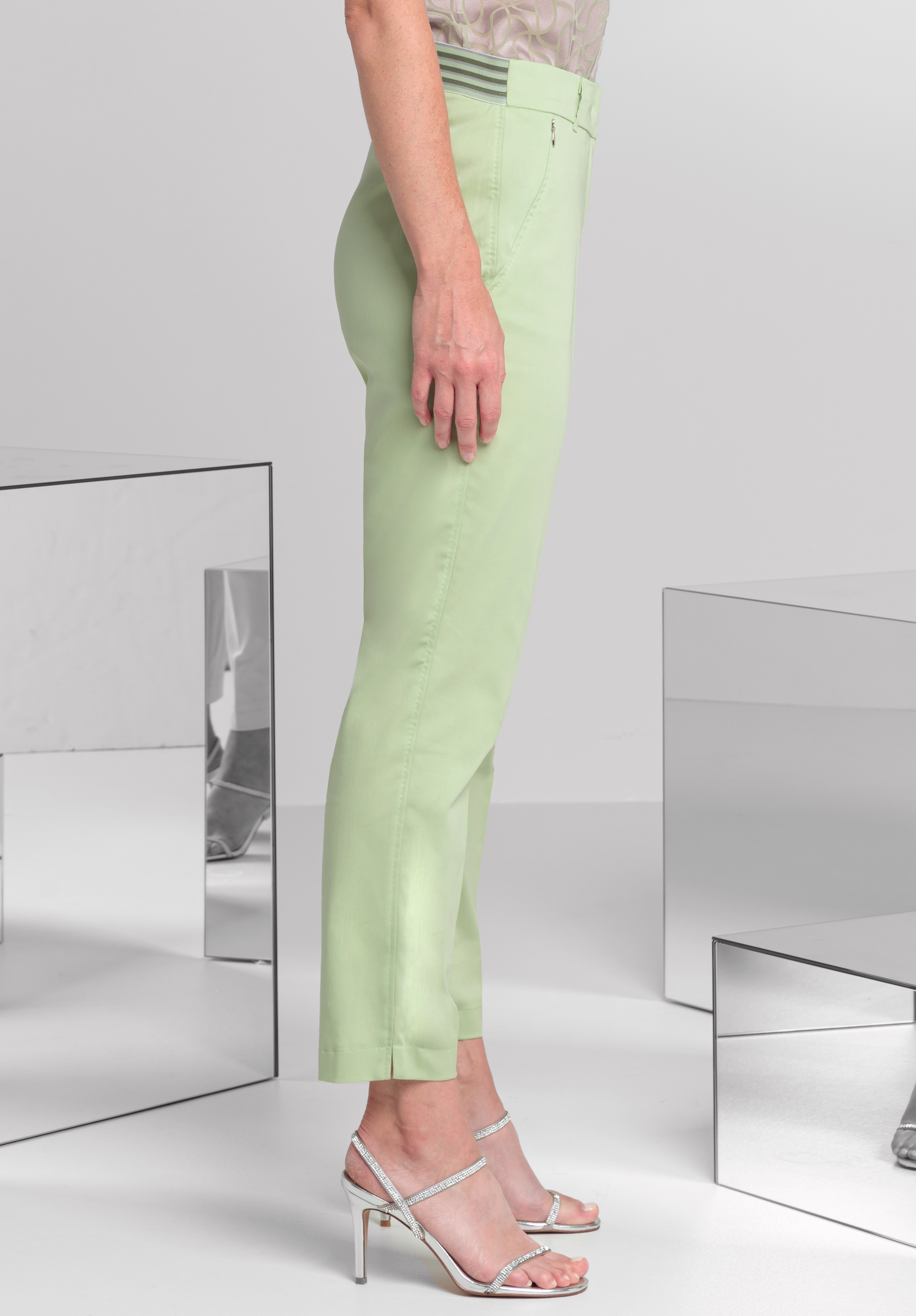 Eine Person steht im Profil und trägt pastellgrüne knöchellange Hosen und silberne Sandalen mit hohen Absätzen vor einem minimalistischen Hintergrund mit spiegelnden Oberflächen.