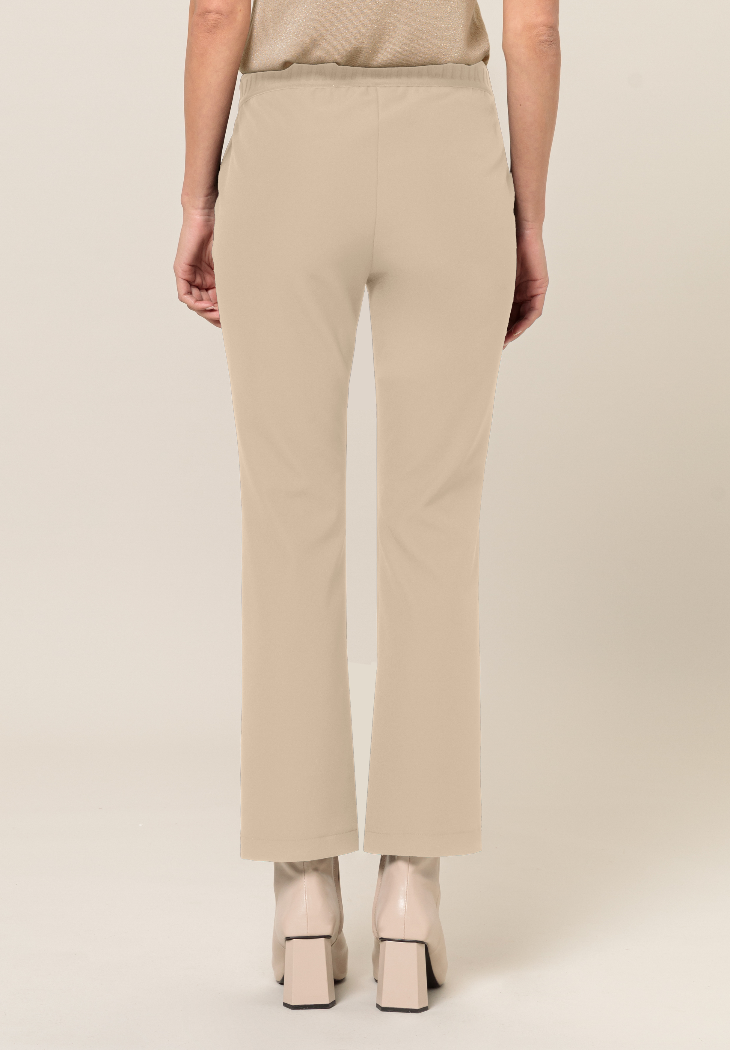 Eine Person steht mit abgewandtem Gesicht da und trägt beige Hosen mit geradem Bein und cremefarbene Stiefel mit quadratischer Spitze und Absätzen vor einem schlichten hellen Hintergrund.