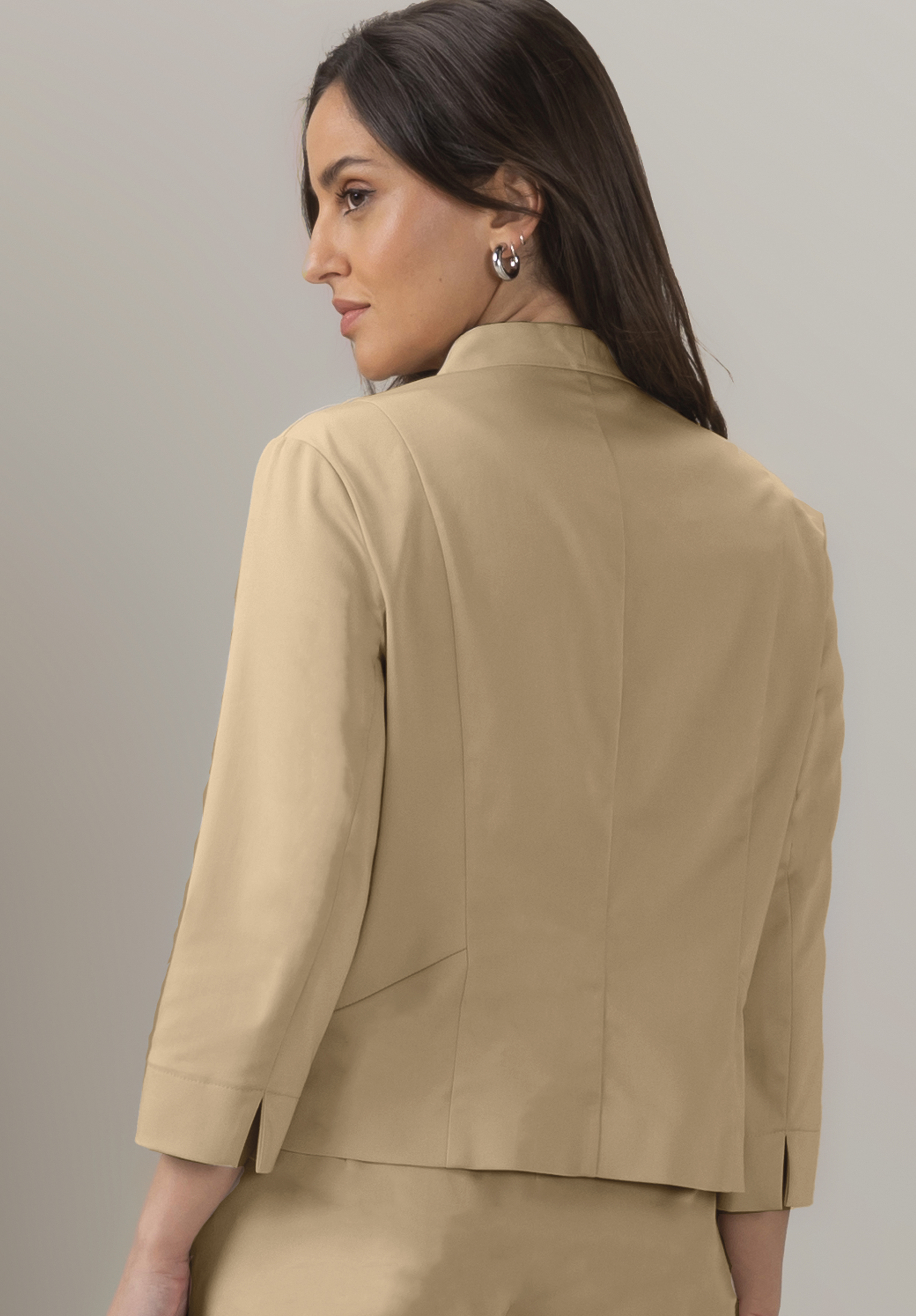 Eine Frau mit langen dunklen Haaren, einem beigen Blazer mit Dreiviertelärmeln, ist von hinten zu sehen und blickt über ihre Schulter. Sie trägt silberne Creolen und hat einen neutralen Gesichtsausdruck.