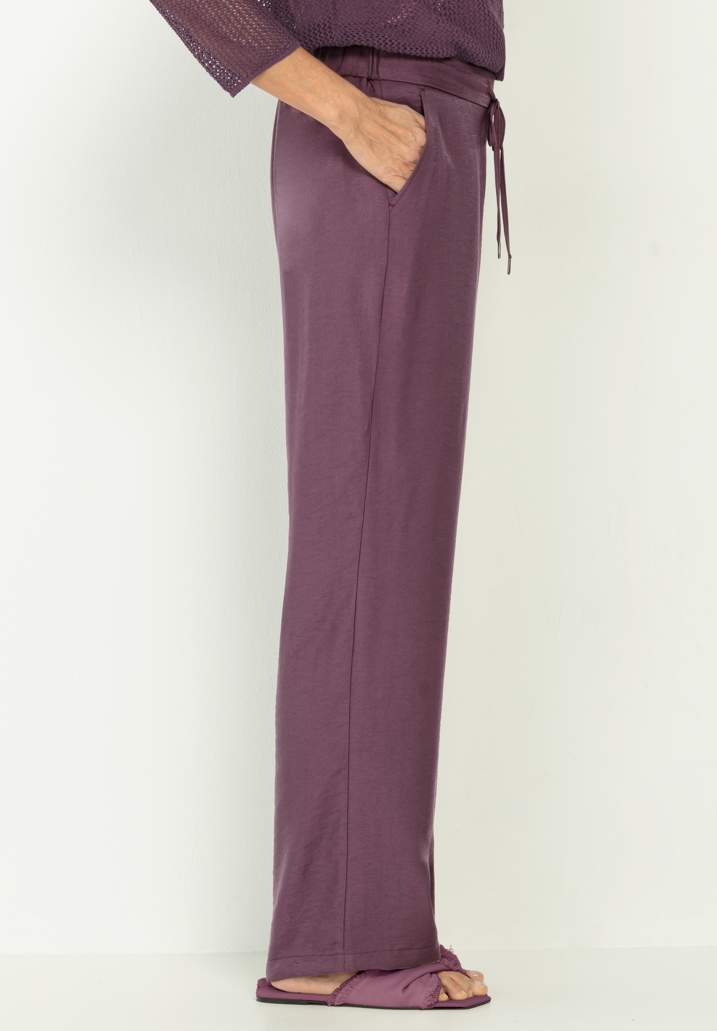 Eine Person steht seitlich und trägt eine weite, lila Hose mit Kordelzug und offene Sandalen, eine Hand in der Tasche, vor einem schlichten weißen Hintergrund.