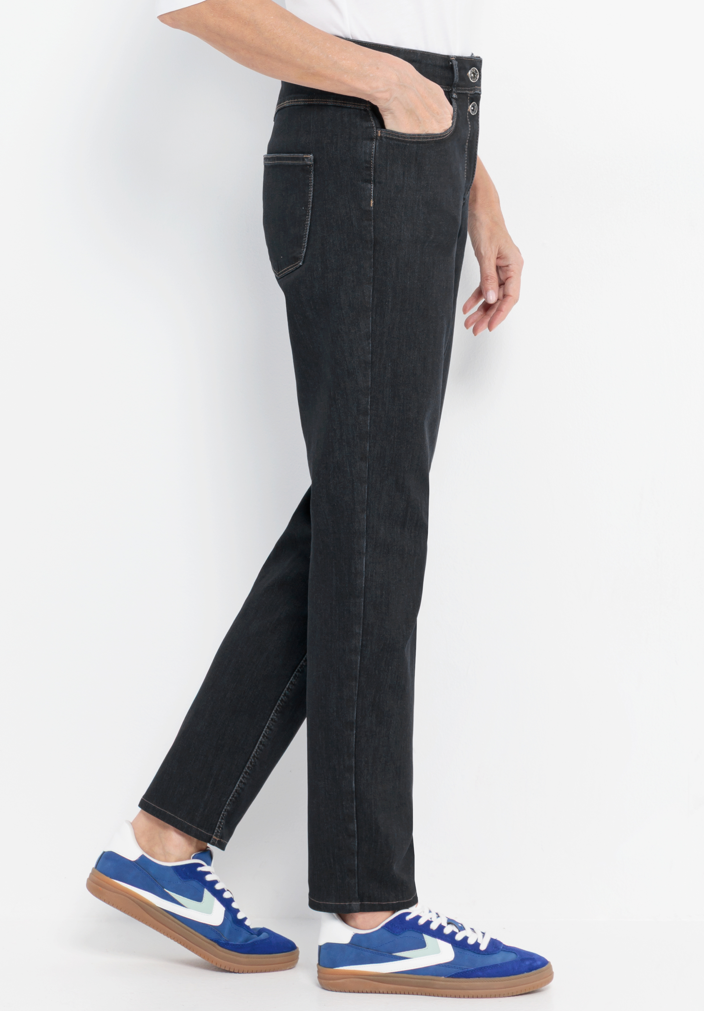 Eine Person, die schwarze Jeans und blau-weiße Turnschuhe trägt, steht seitlich mit einer Hand in der Hosentasche vor einem schlichten weißen Hintergrund.