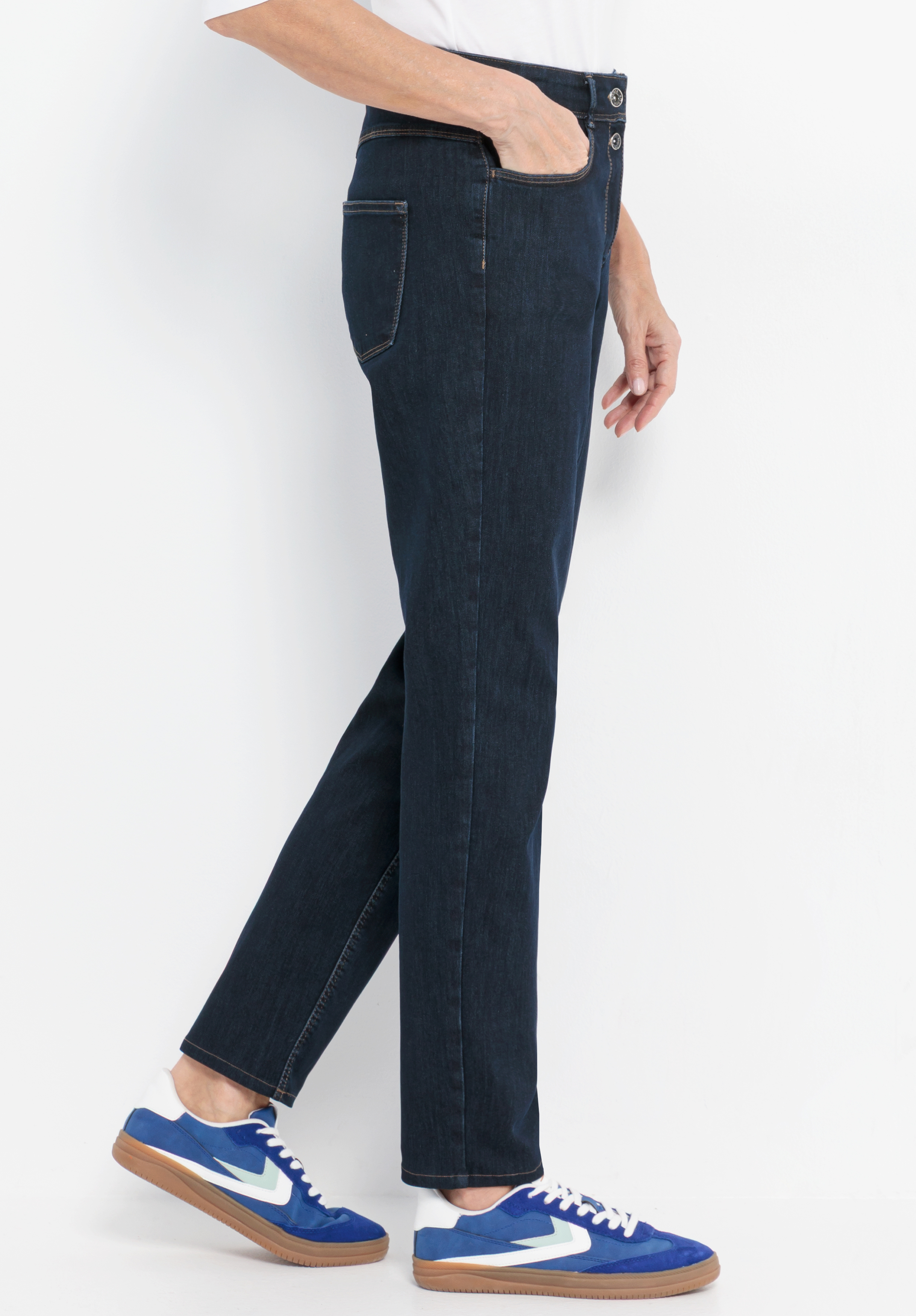 Eine Person, die dunkelblaue Jeans und blaue Turnschuhe mit weißen Sohlen trägt, steht vor einem weißen Hintergrund, eine Hand in der Hosentasche, von der Seite gesehen.